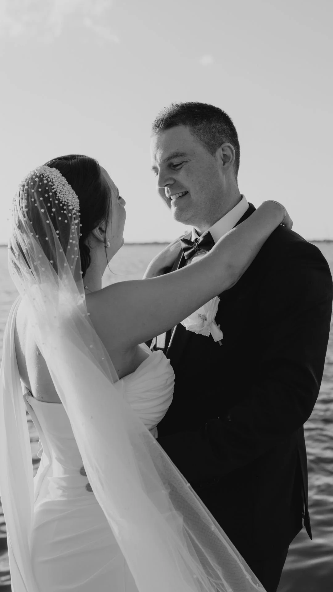A black and white photo of a bride and groom embracing outdoors by the water, smiling at each other, with the bride wearing a wedding gown and veil and the groom in a tuxedo.