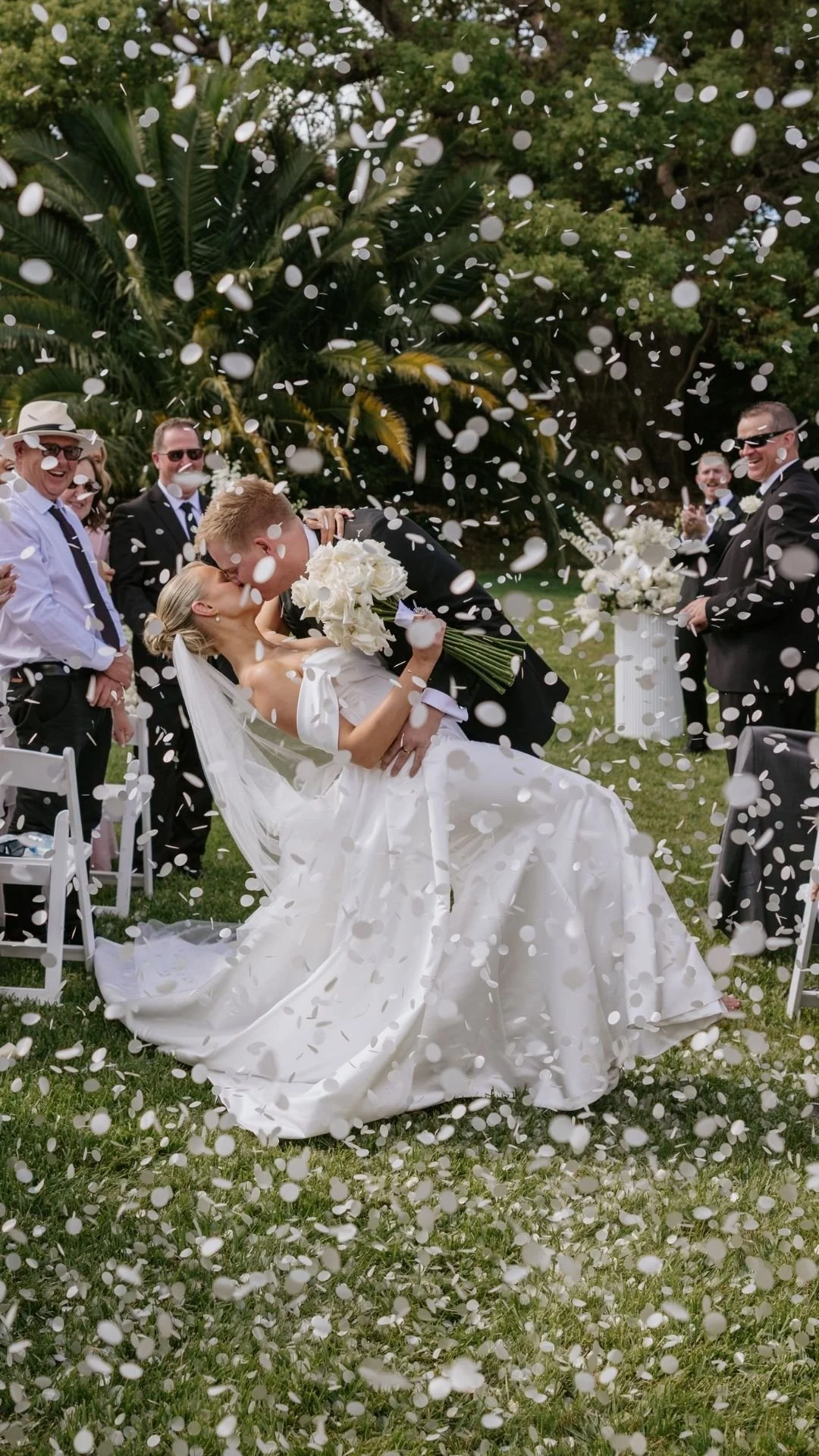 A newlywed couple kisses while being showered with confetti during their wedding ceremony outdoors, with family and friends celebrating around them.