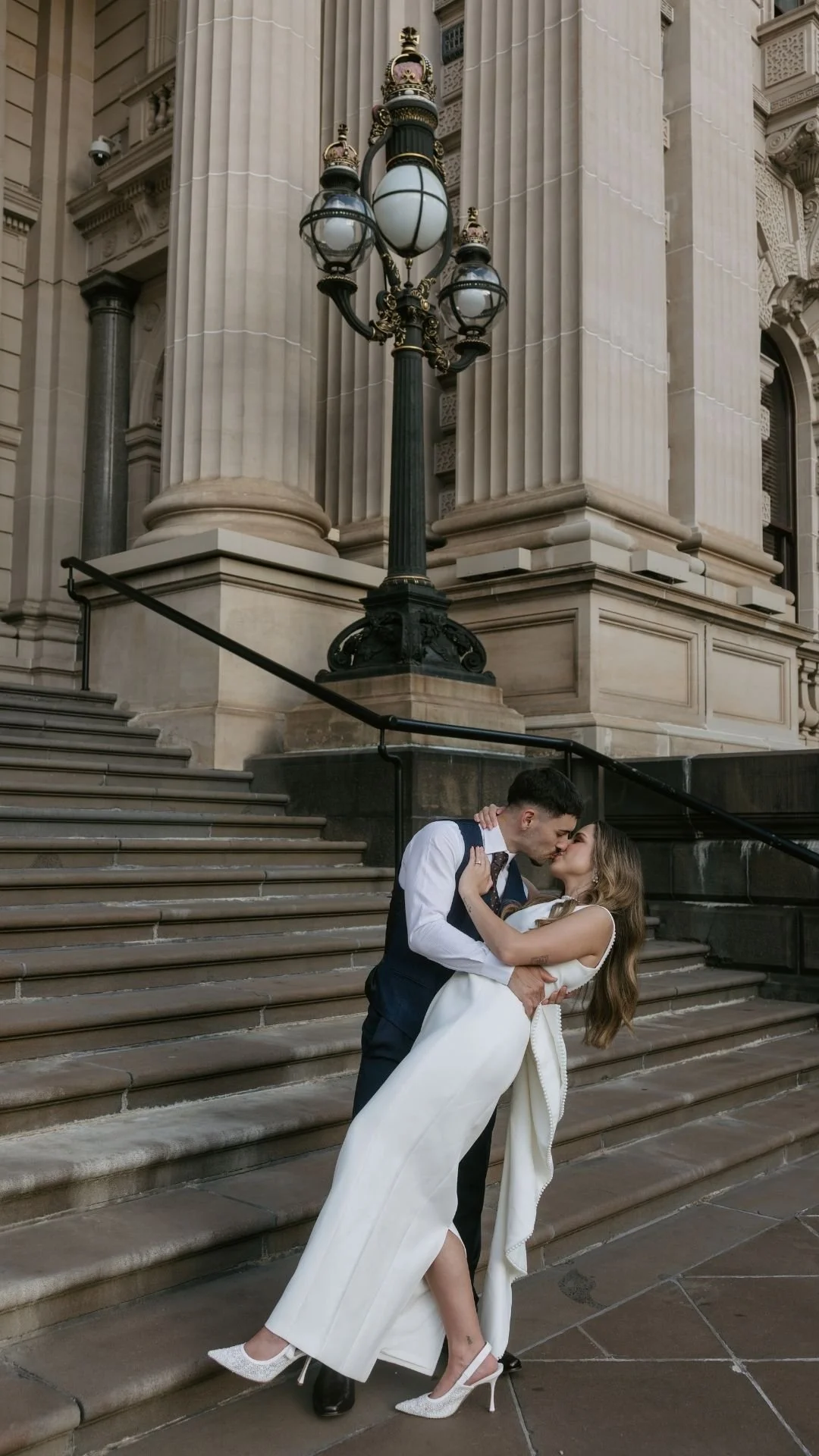 A couple in formal attire sharing a kiss on the steps in front of a building with large columns and vintage street lamps.