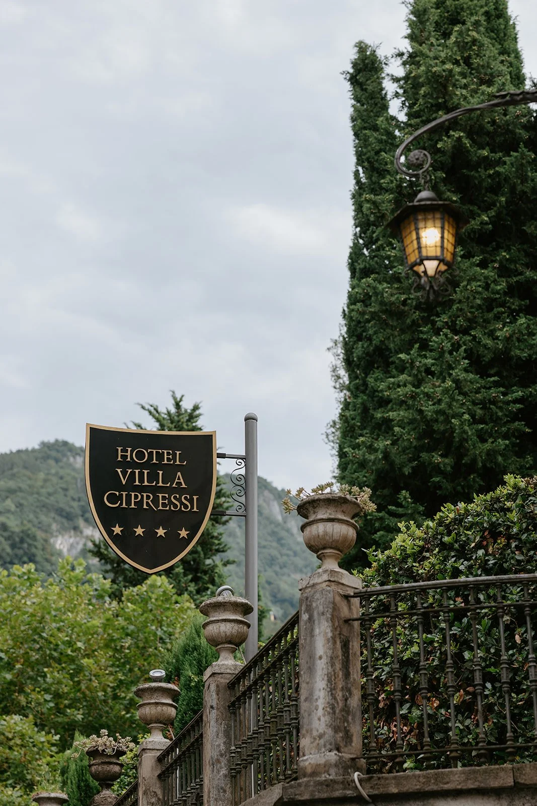Sign for Hotel Villa Cipressi with four-star rating, ornate lamp post, stone railing, lush greenery, and cloudy sky in the background.
