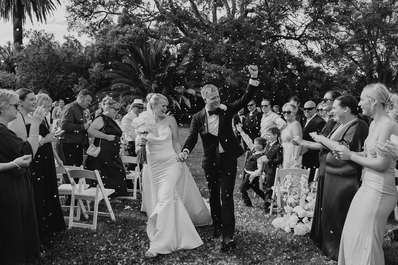 A black-and-white photo of a wedding celebration outdoors, showing the bride and groom walking hand in hand through a crowd of smiling guests, with confetti in the air and trees in the background.