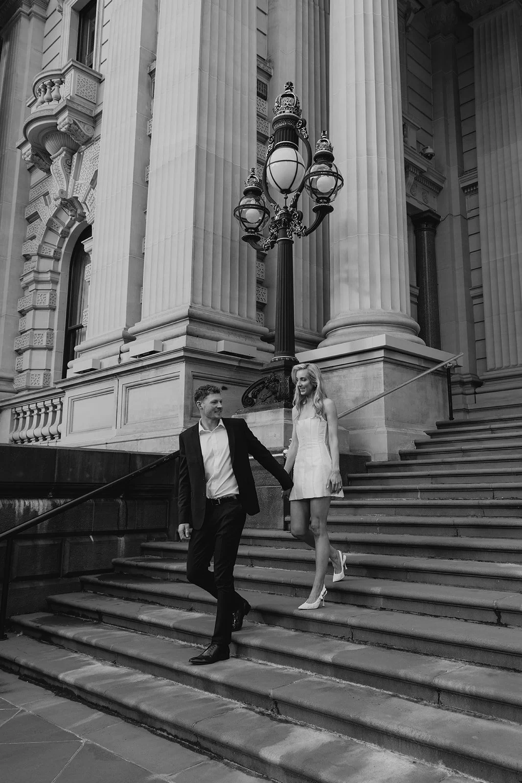 A man in a suit and a woman in a dress are walking down stairs outside a large classical building with tall columns and ornate architectural details, holding hands and smiling.