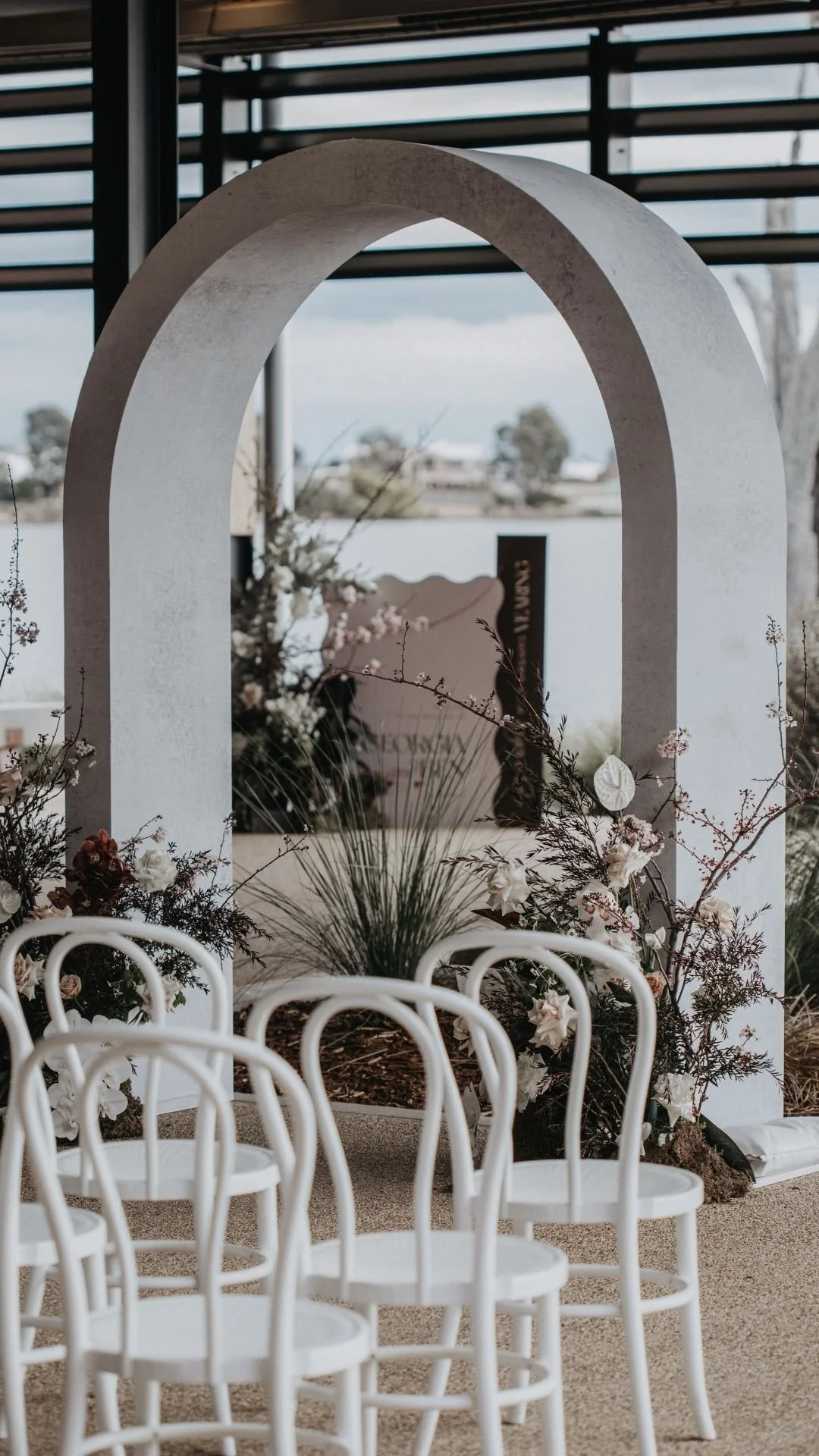 Wedding ceremony setup with white chairs, floral arrangements, and a large decorative arch outdoors near a body of water.