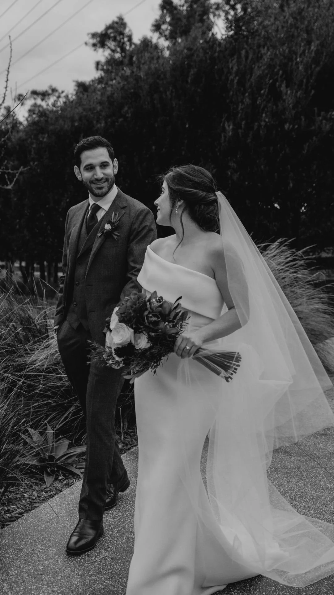 Black and white photo of a bride and groom walking outdoors, the bride holding a bouquet and looking at the groom, both dressed in wedding attire, with trees and plants in the background.