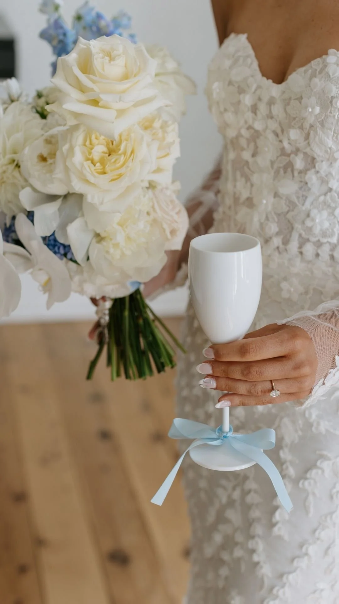 A bride holding a bouquet of white and light purple flowers with one hand, and a white champagne flute with a light blue ribbon tied around its stem in the other hand, wearing a lace wedding dress.