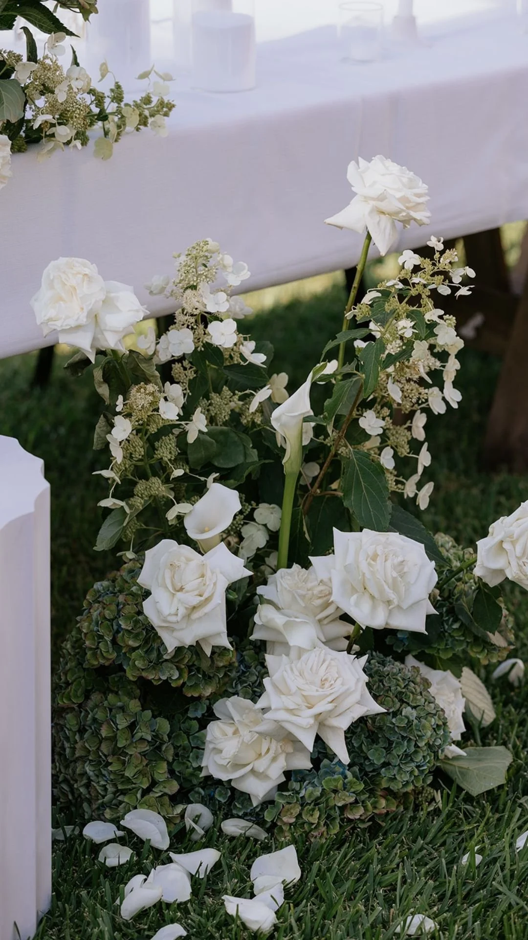White flower arrangement with roses, calla lilies, hydrangeas, and greenery on green grass, part of a decorated table setup for an event.