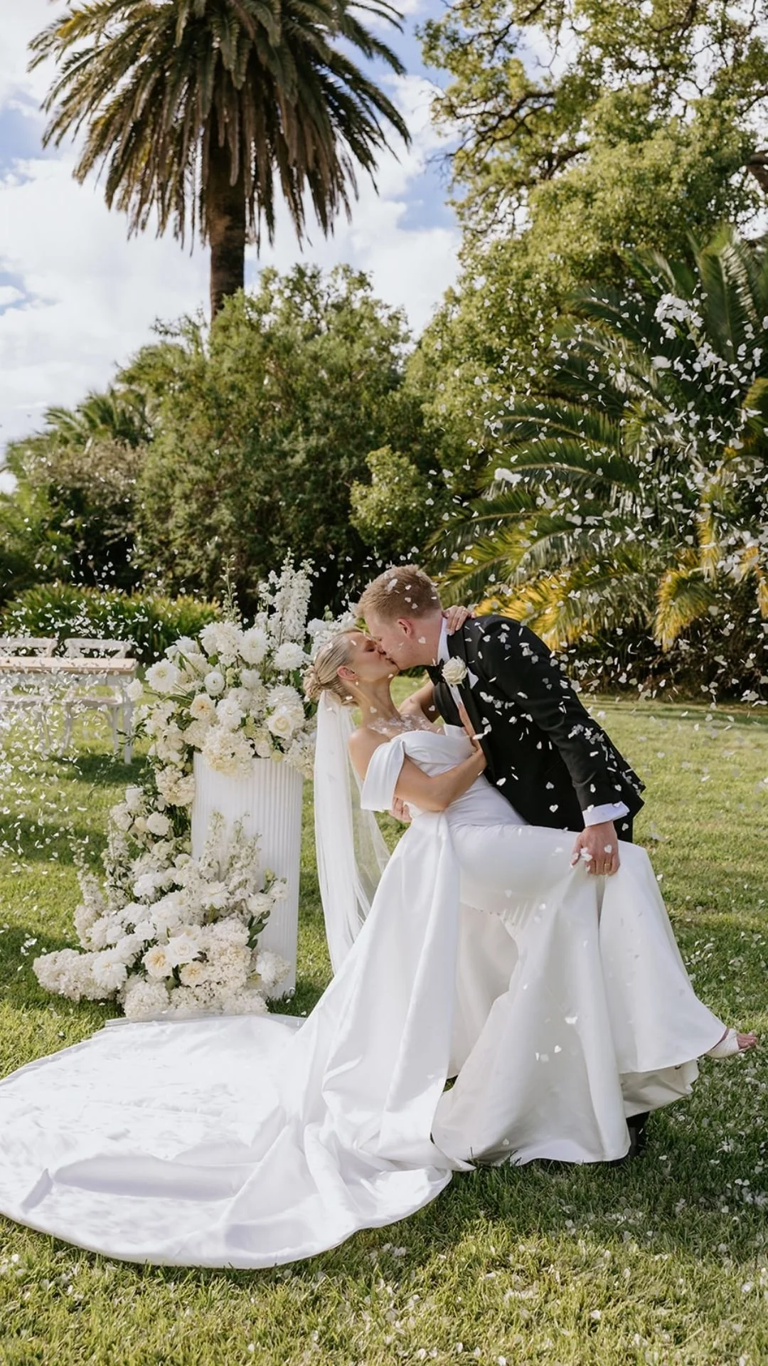 Bride and groom sharing a kiss at their wedding outdoors, with a large floral arrangement and lush trees in the background.