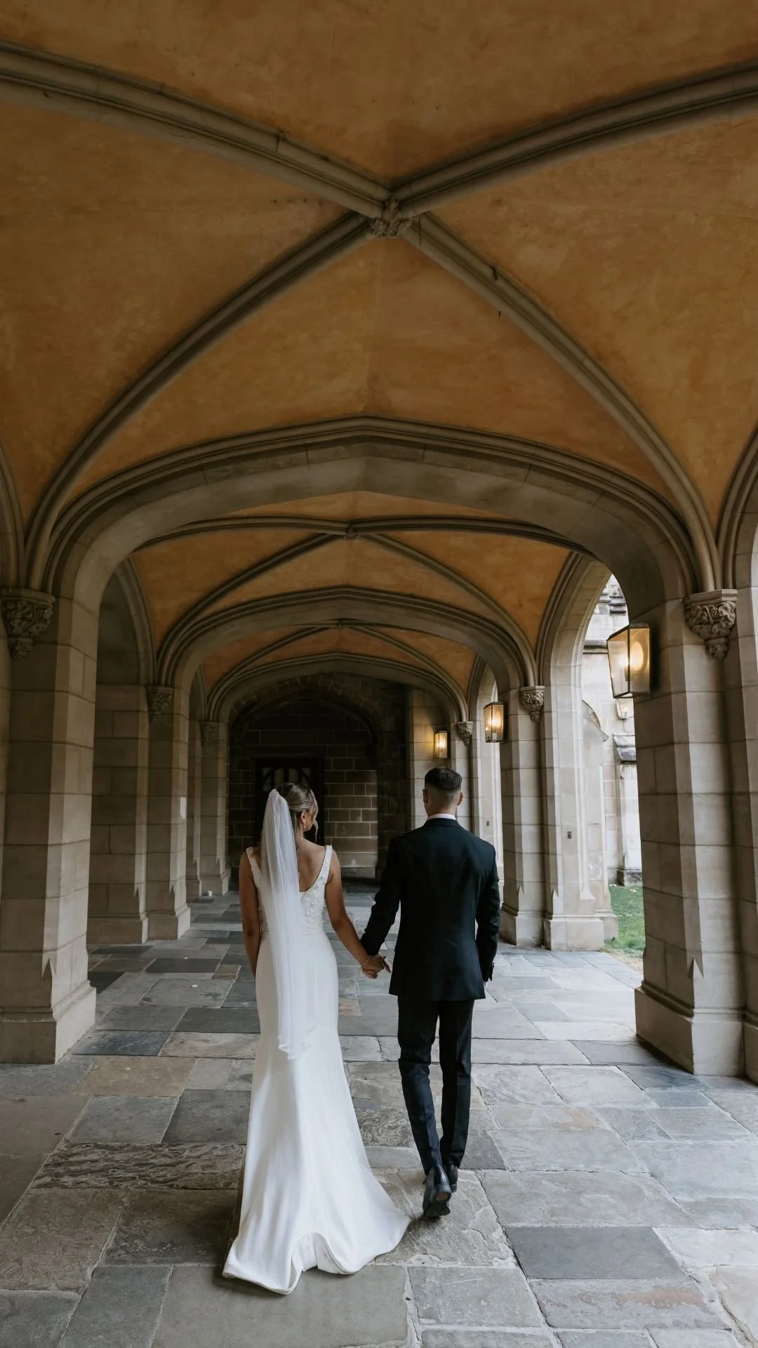 A bride and groom holding hands walking through an arched stone hallway, with the bride in a white gown and veil and the groom in a black suit.