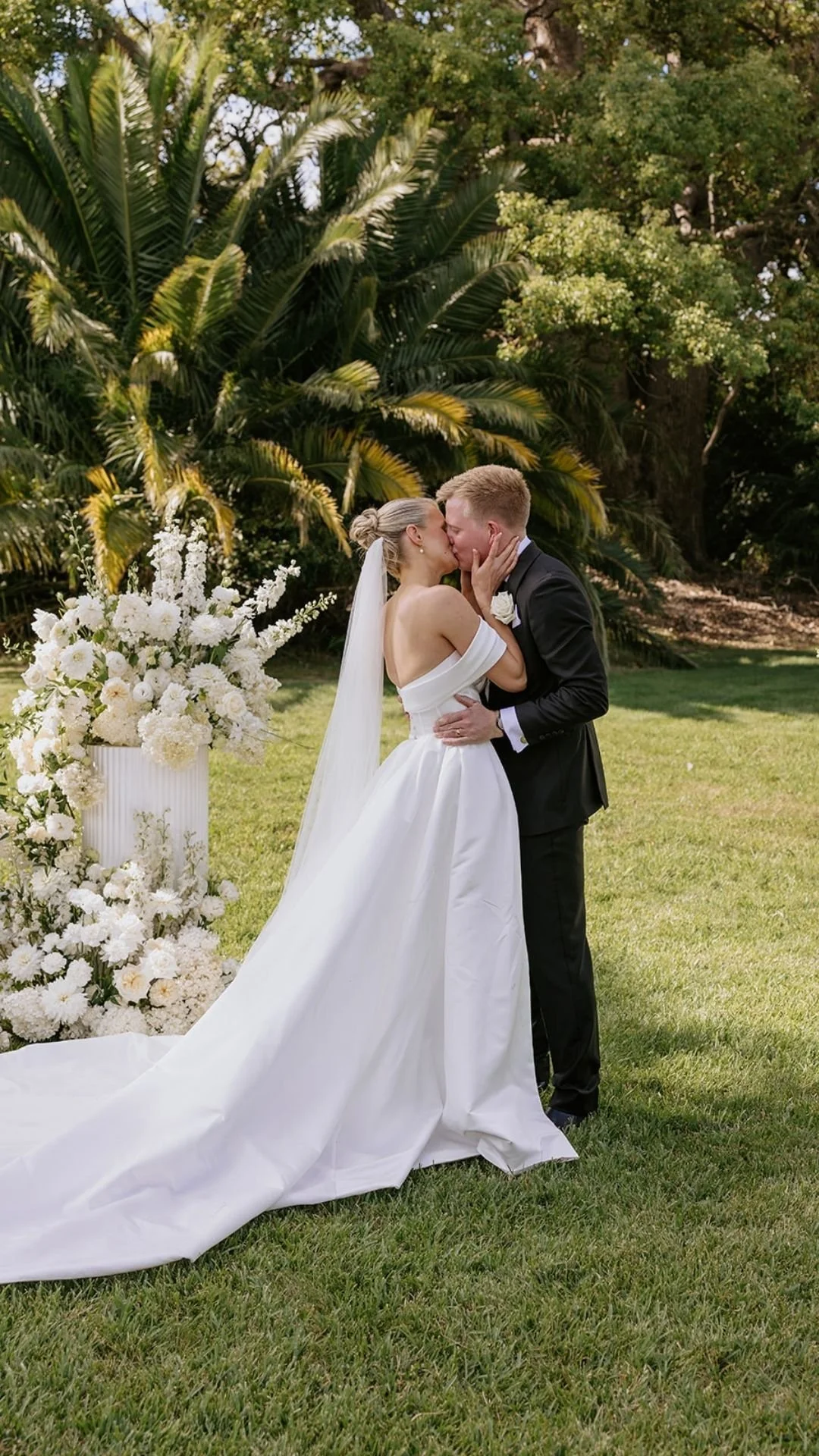 A bride and groom kissing outdoors on their wedding day, in front of a large white floral arrangement with greenery backgrounds.