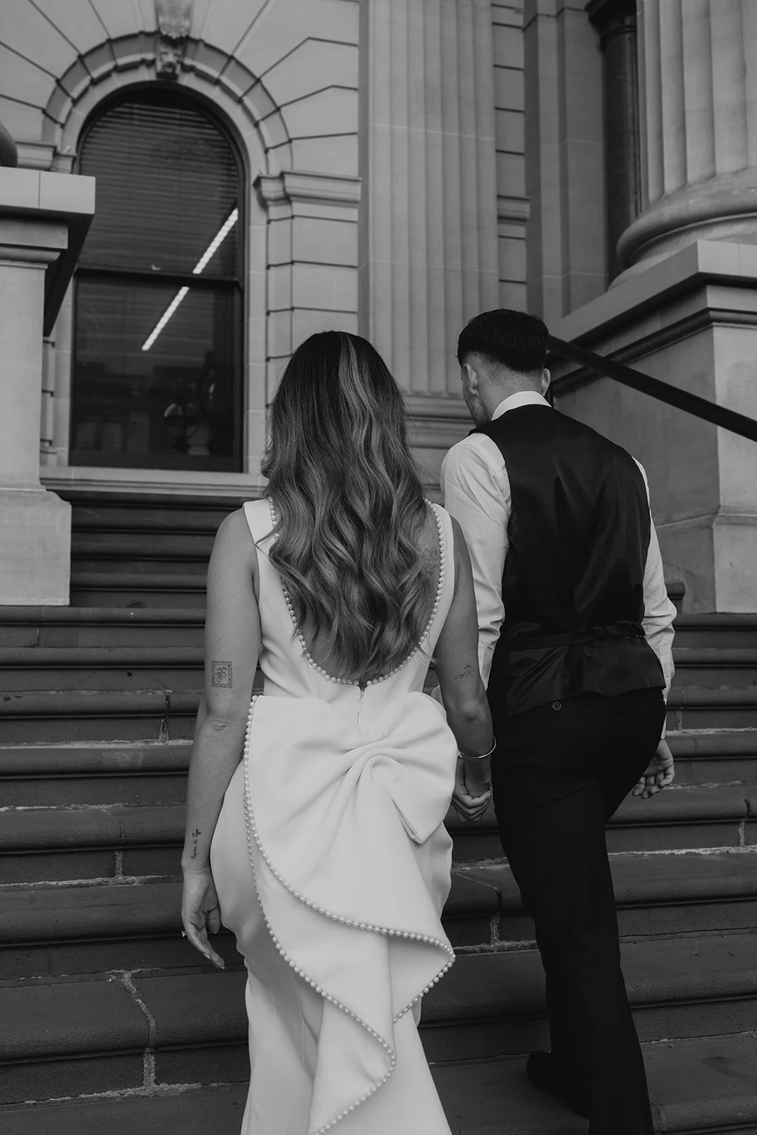 A couple dressed in formal attire walking up the steps of a large, historic building with elaborate stone architecture.