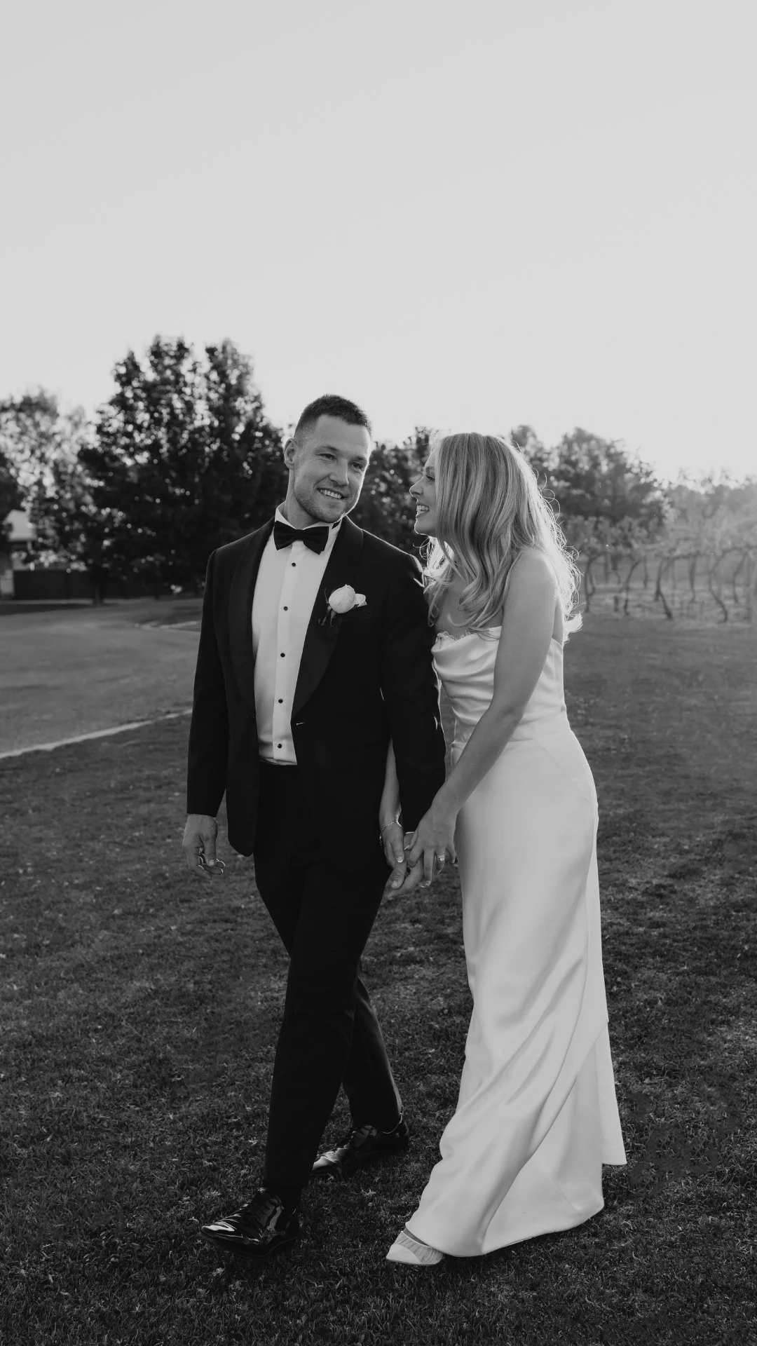Morrisons Winery happy couple in wedding attire holding hands and smiling outdoors with Echuca Moama wedding photographer love by shae in a grassy area with trees in the background in black and white.