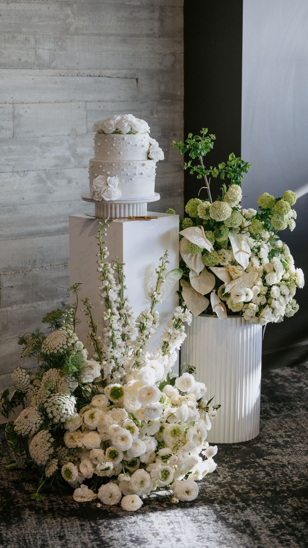 A white wedding cake decorated with white roses and small pearl-like embellishments, displayed on a pedestal, surrounded by white floral arrangements including roses, hydrangeas, and other white flowers, against a grey textured wall.