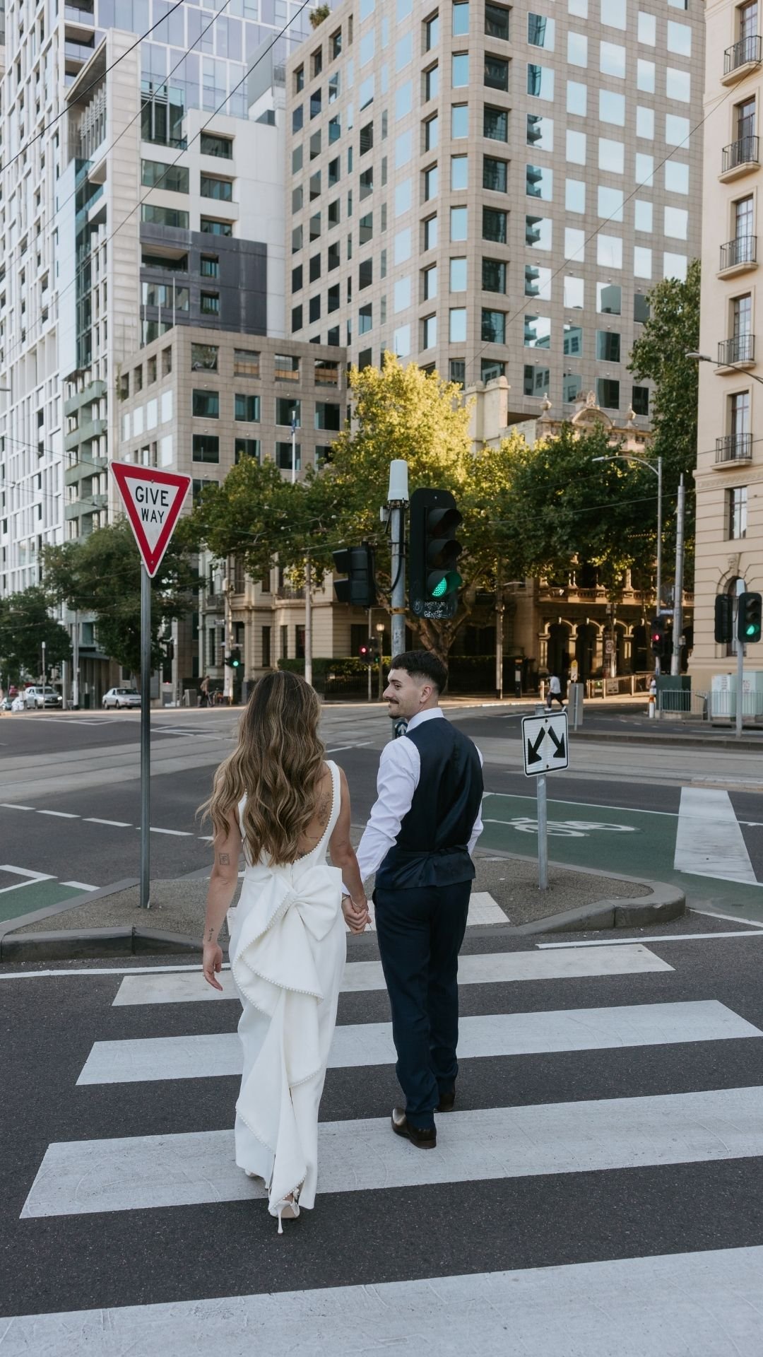 A young couple, dressed in formal attire, holding hands and crossing a city street at a crosswalk, with tall buildings and trees in the background.