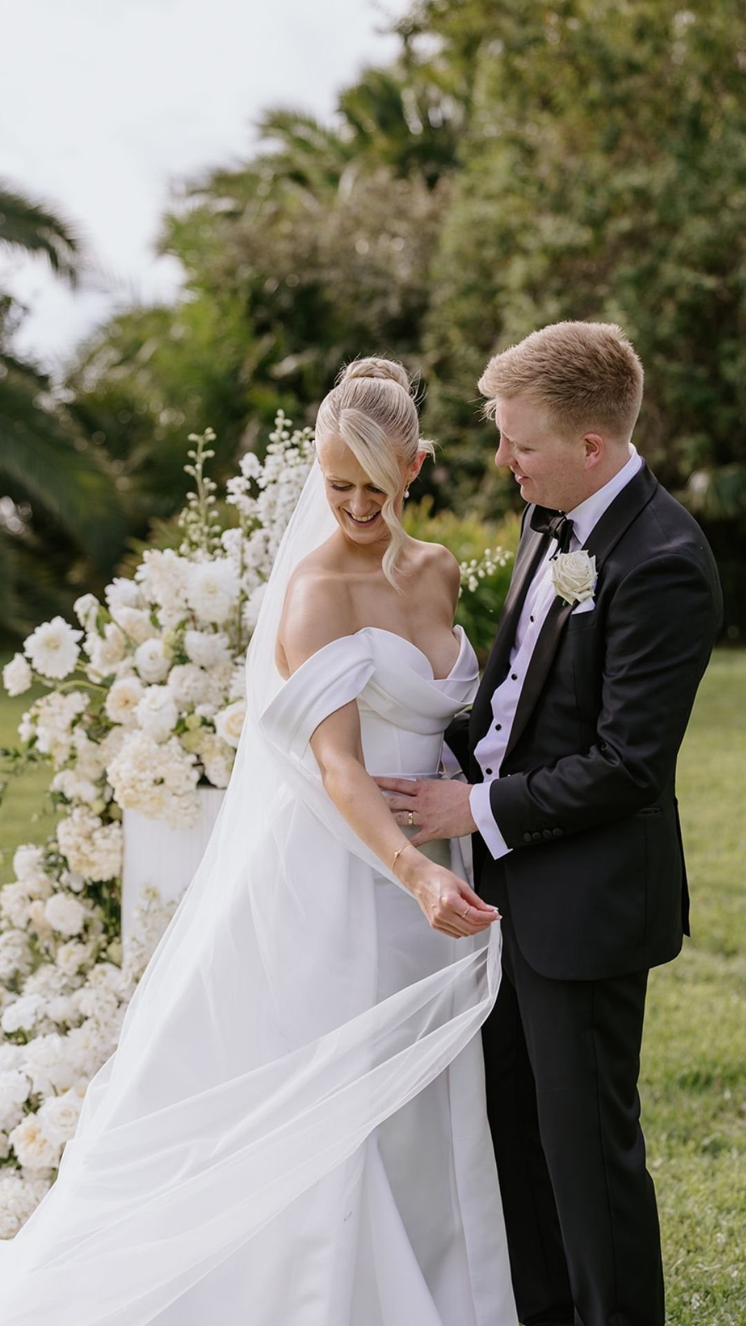 A bride and groom sharing a moment during their outdoor wedding, with the bride in a white gown and the groom in a black tuxedo, standing in front of a floral arrangement of white flowers on a grass lawn with trees in the background.