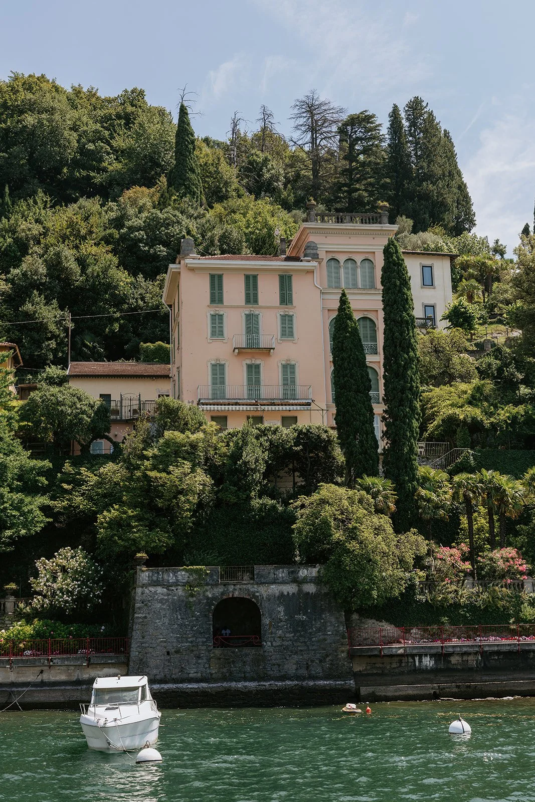 A pink multi-story house with green shutters and a small balcony, situated on a hillside covered with lush green trees, overlooking a body of water with a small white boat and buoys.