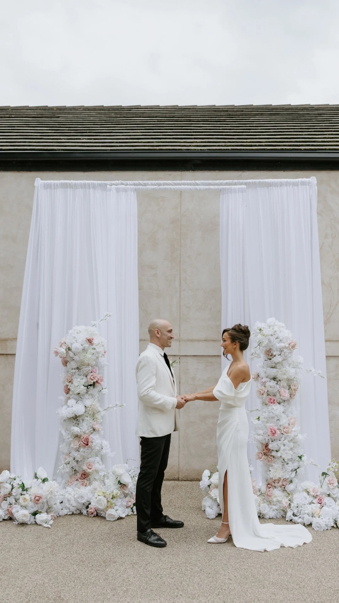 A couple getting married outdoors, holding hands and facing each other, with a floral wedding backdrop of white and pink roses and white drapery.