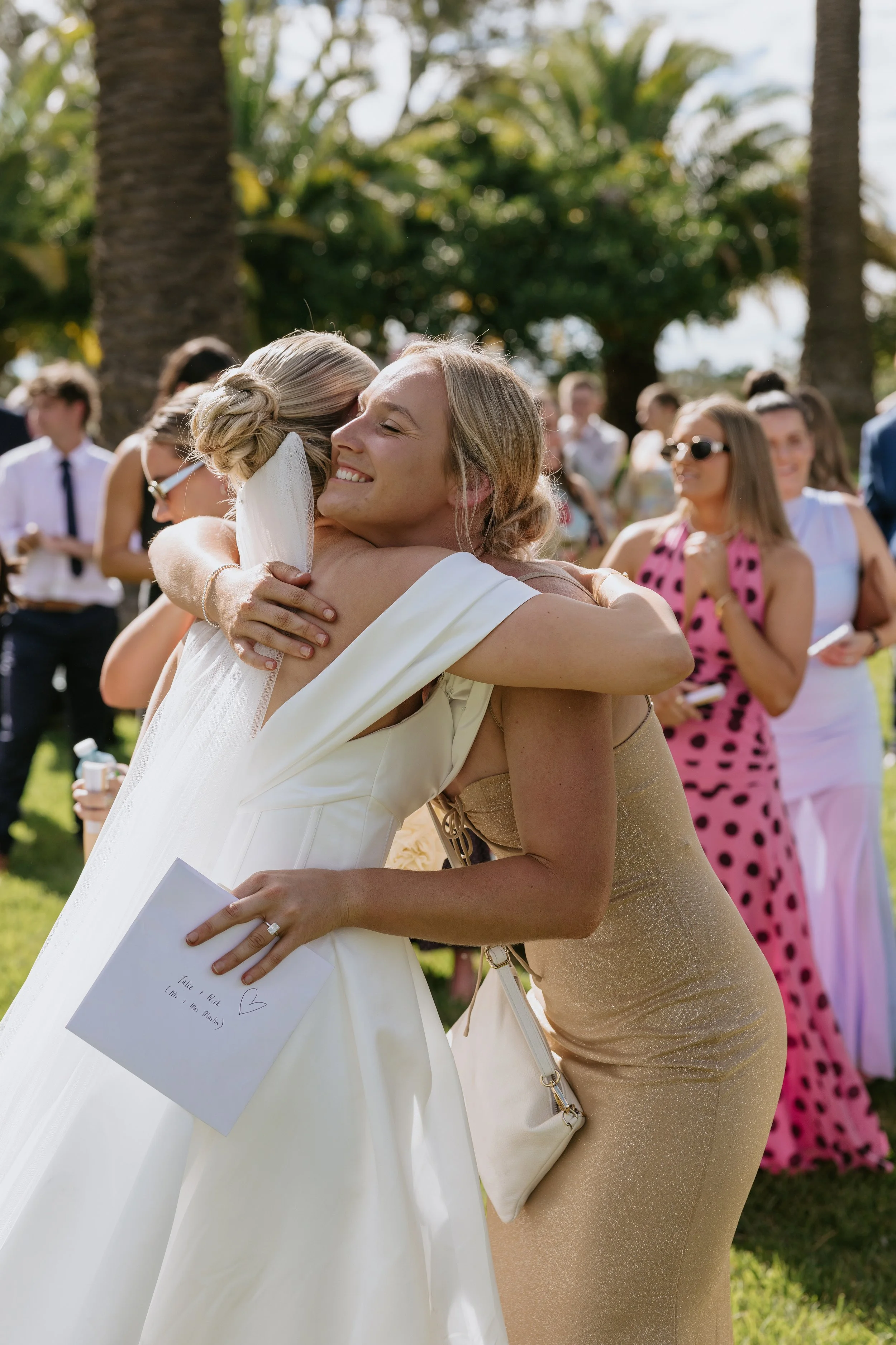 Two women hugging at an outdoor event with other people in the background, lush trees, and bright sunlight.
