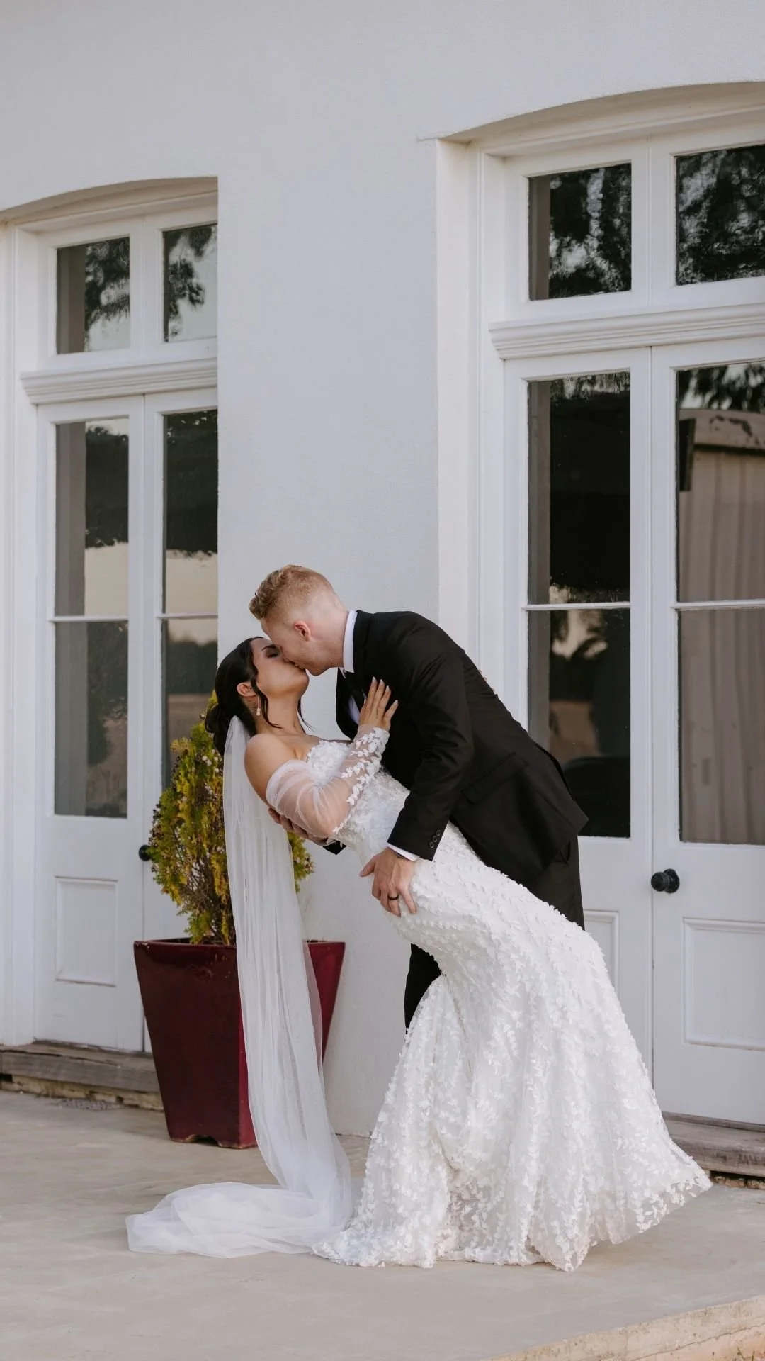 A newlywed couple kissing outside a building with white walls and windows; the groom in a black tuxedo dips the bride in her white wedding dress with lace sleeves.