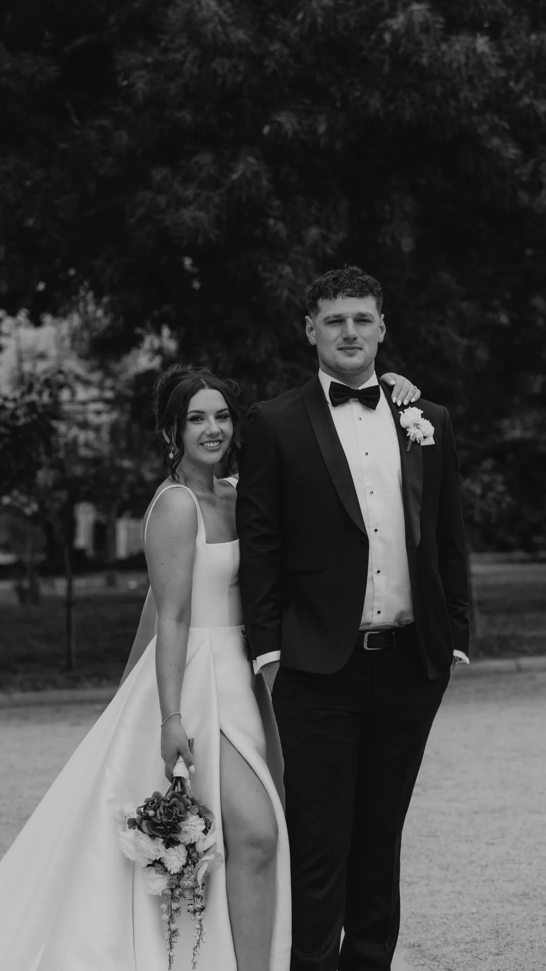 A black and white photo of a bride and groom standing outdoors, with the bride smiling and holding a bouquet, and the groom in a tuxedo, arm around her shoulders, in front of trees.