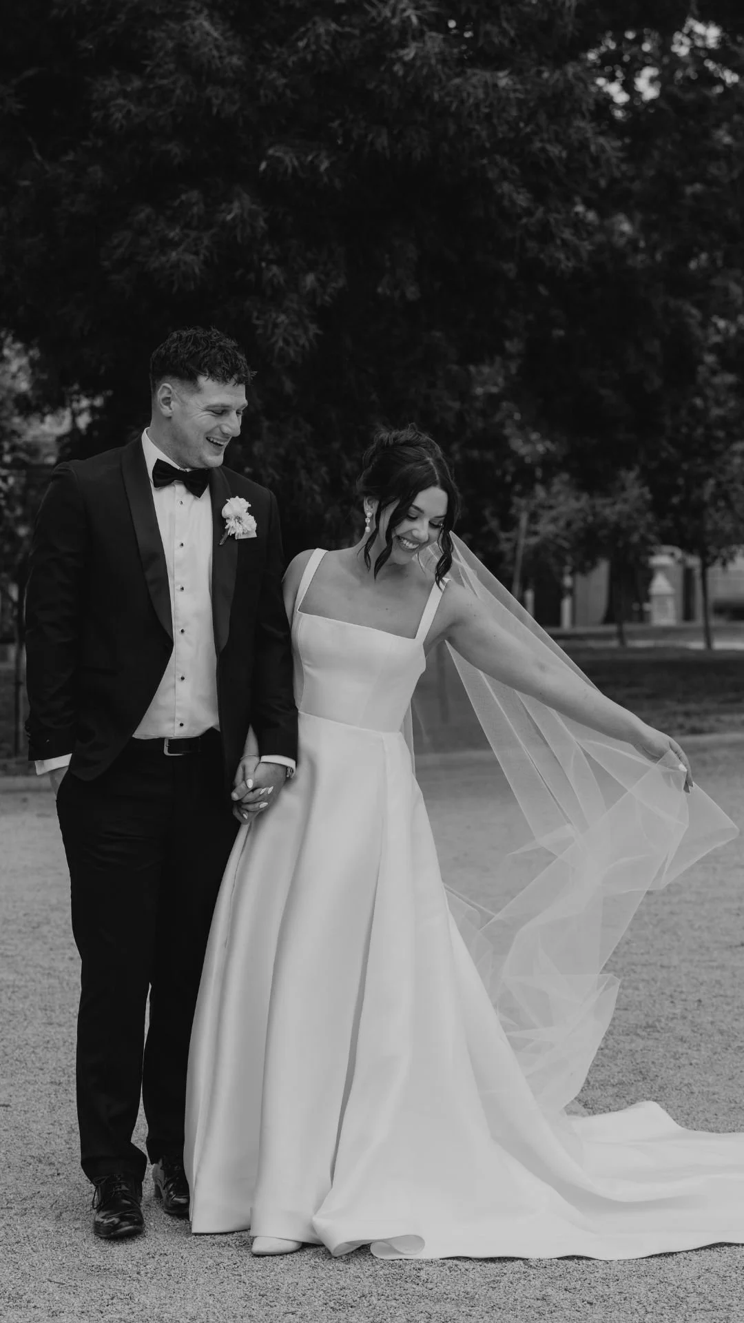 Black and white photo of a bride and groom walking hand in hand outdoors, smiling, with the bride holding up her wedding dress train.