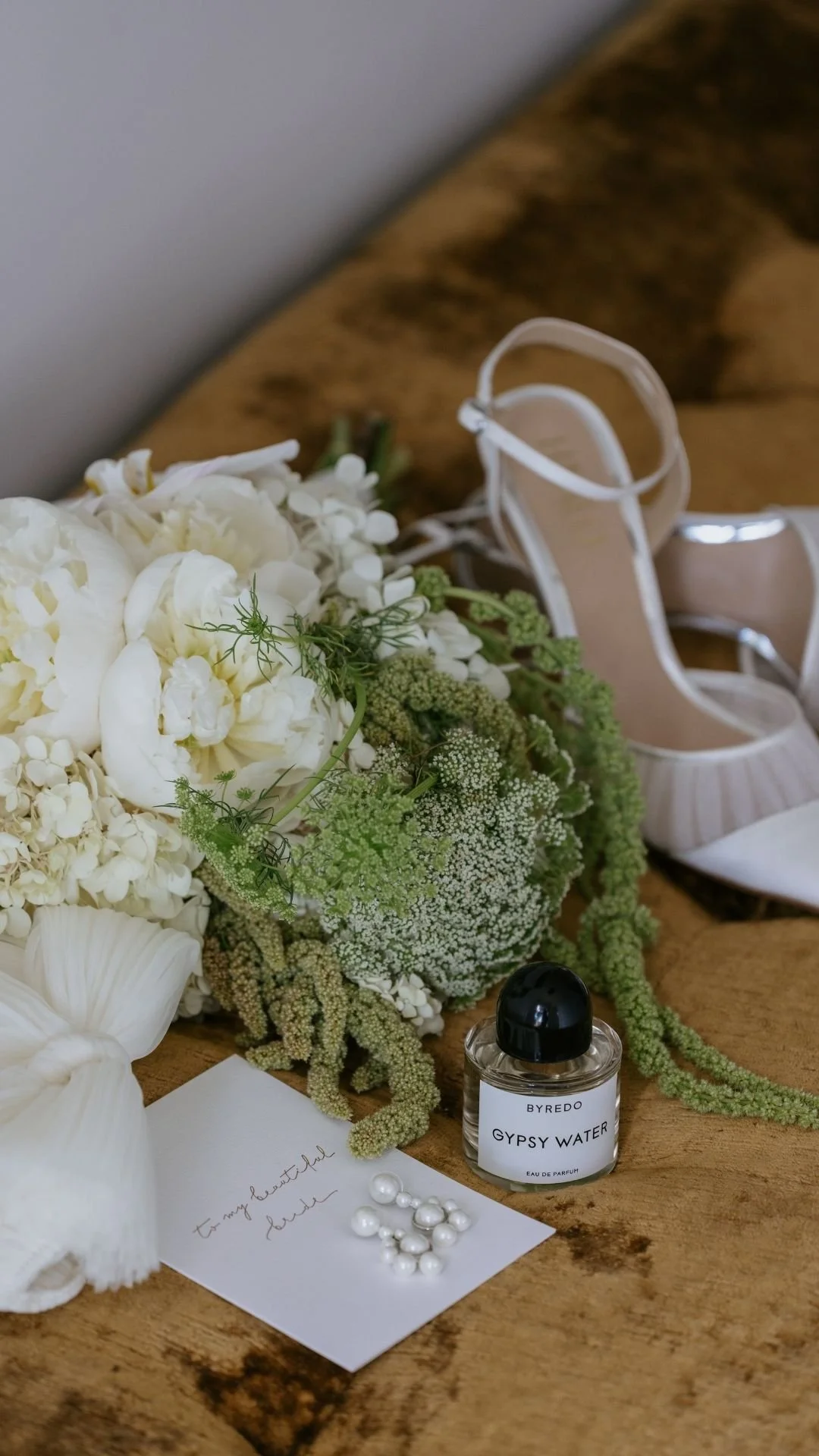 White flower bouquet, perfume bottle labeled 'BYREDO GYPSY WATER', white high-heeled shoe, small bottle of Gypsy Water perfume, handwritten note, pearl embellishments, and a piece of paper with handwritten text placed on a wooden surface.