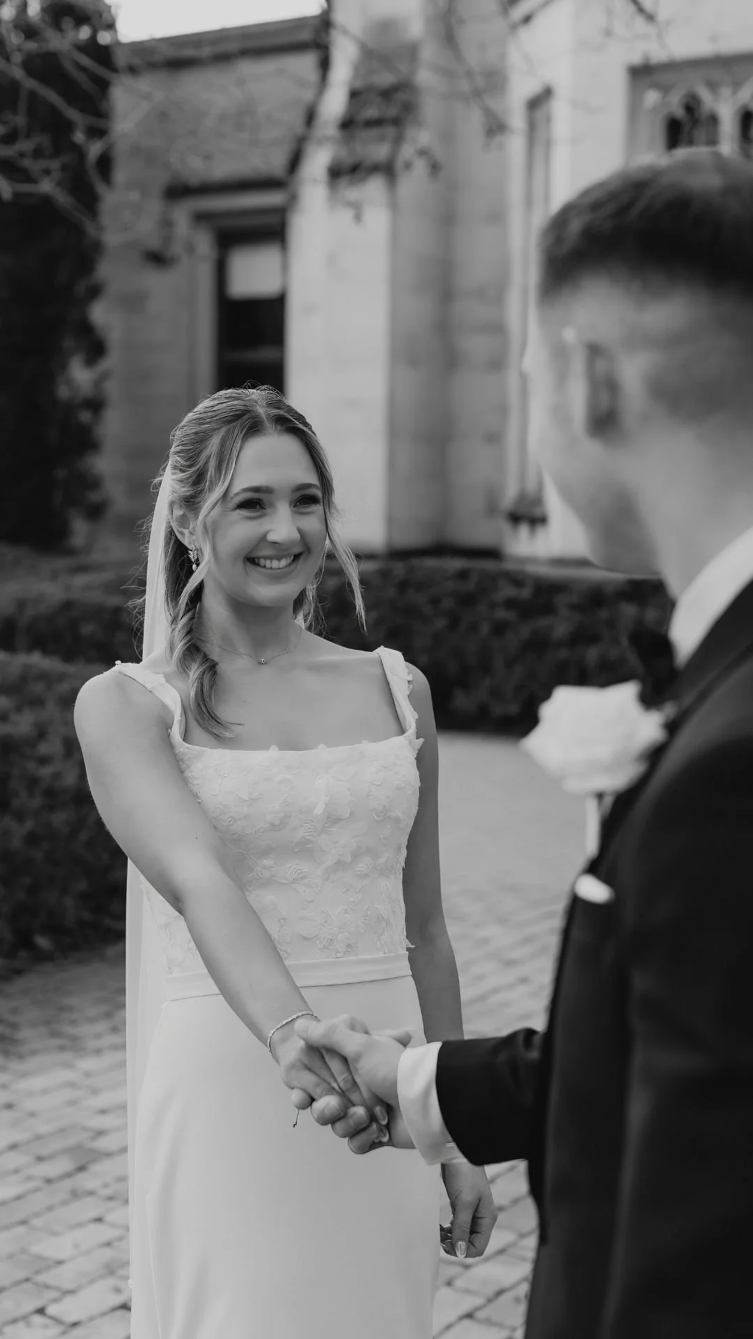 A bride and groom holding hands, standing outdoors in front of a building. The bride is smiling, wearing a white wedding dress with lace details, and has a veil. The groom, in a dark suit, is partially visible, facing the bride.