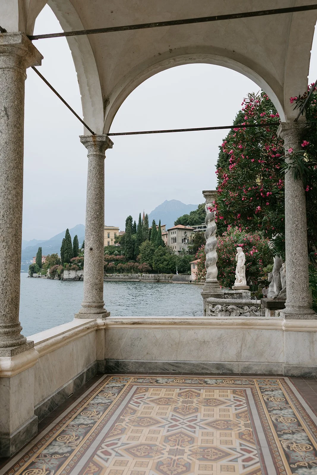 View of a lake with buildings and mountains in the background, framed by a marble balcony with columns and decorative statues, and pink flowering bushes to the right.