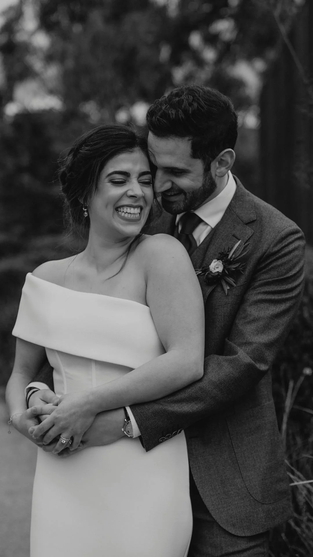Black and white image of a bride and groom laughing and embracing outdoors. The bride wears an off-shoulder wedding dress, and the groom wears a suit with a boutonniere.