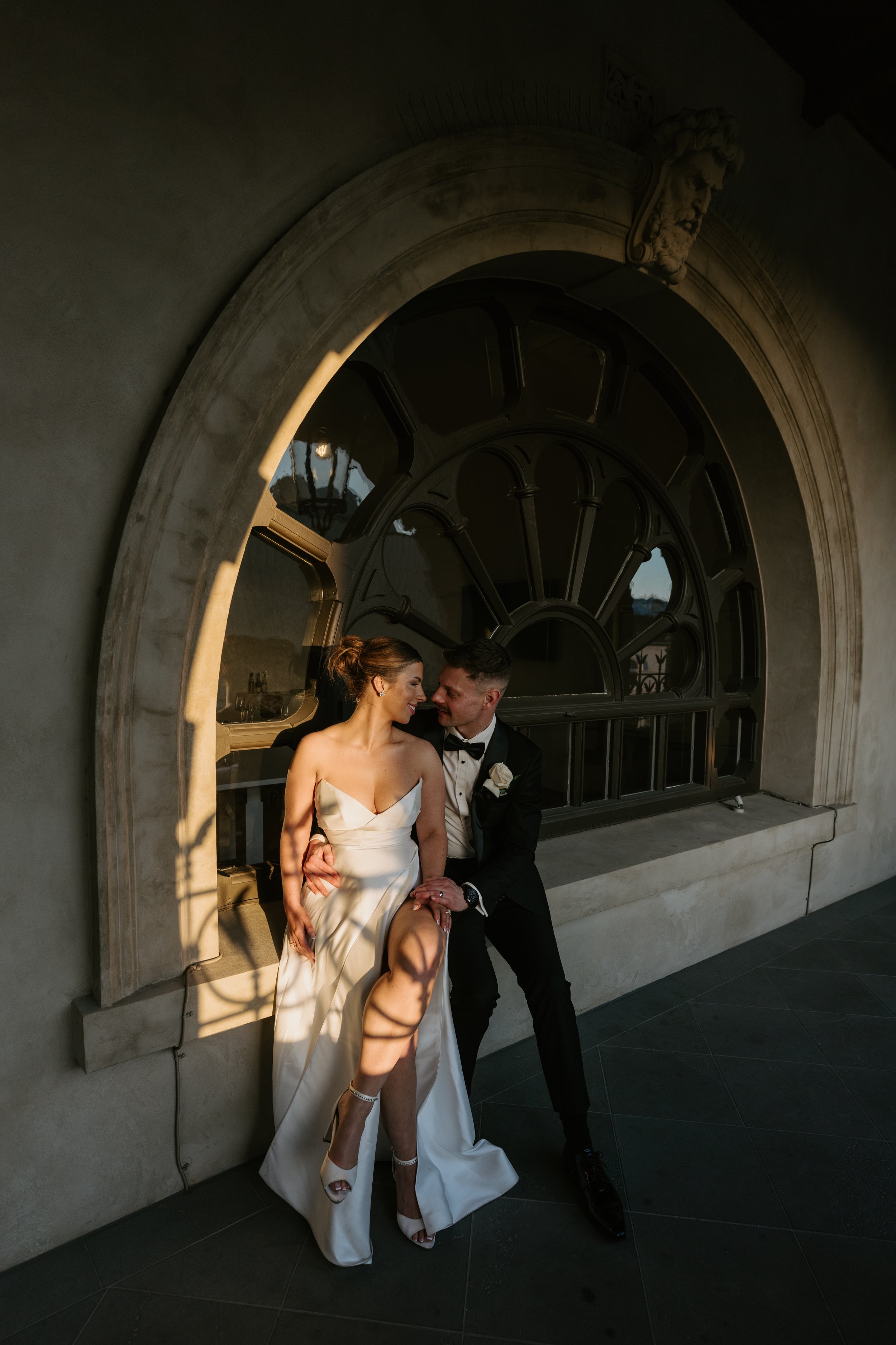 A newlywed couple sits on a stone window sill, sharing a loving moment. The bride wears a white strapless wedding gown and high heels, and the groom is dressed in a black tuxedo with a white boutonniere. They are both smiling and leaning toward each other. The setting is outdoors, with the sunlight casting shadows through an arched window with a decorative lion's head sculpture above, creating a romantic atmosphere.