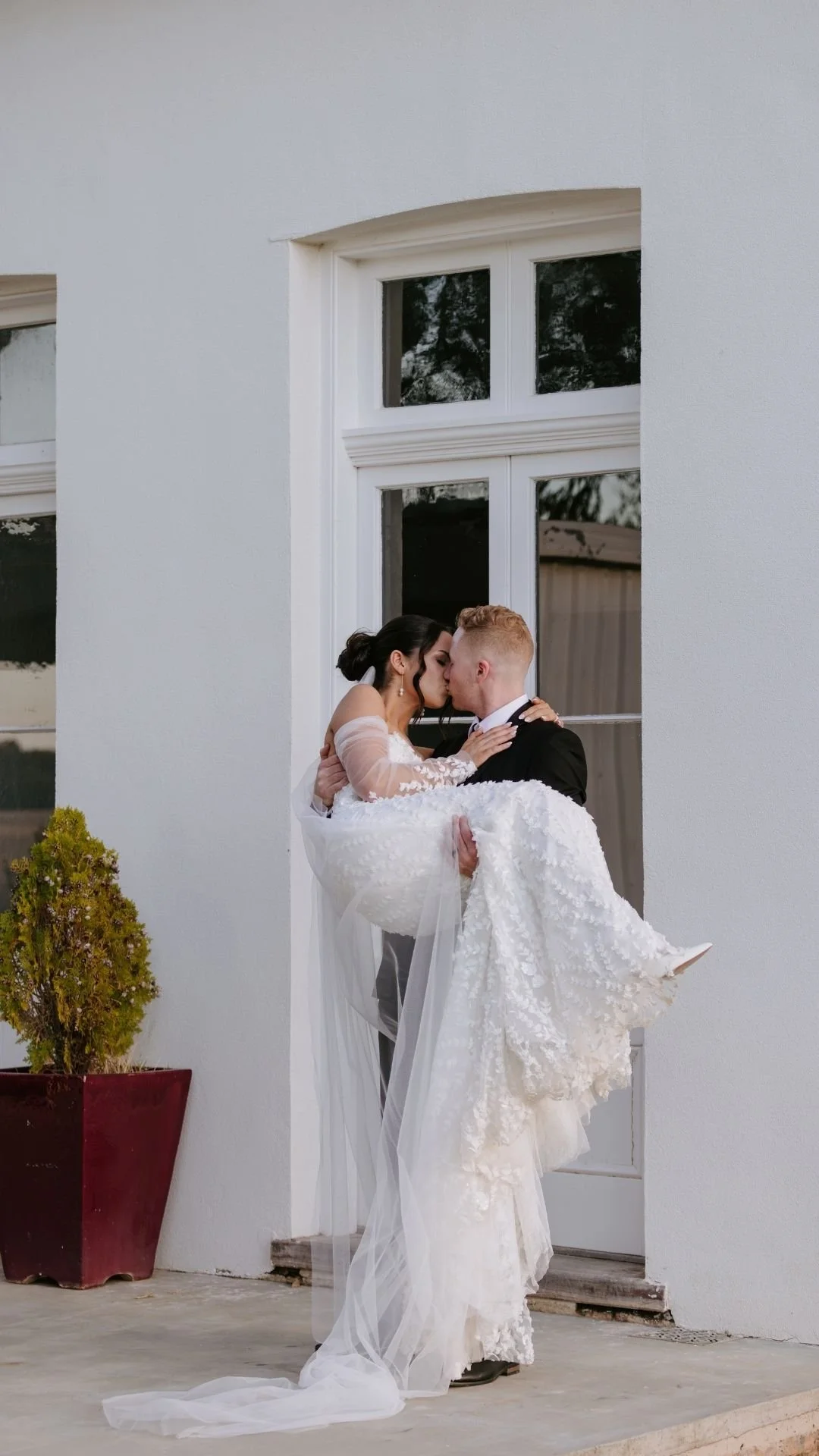 A bride in a white wedding dress being carried by a groom in a black tuxedo, both sharing a kiss outside in front of a white wall with large windows.