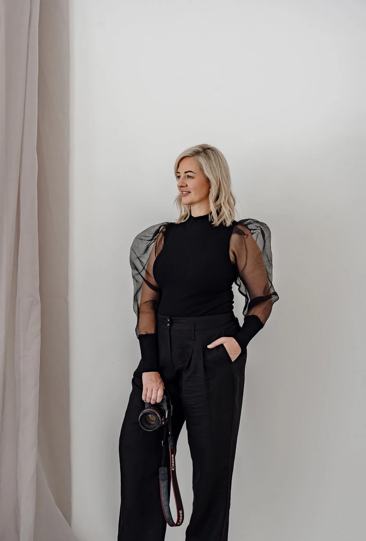 A woman with blonde hair wearing a black top with sheer puffed sleeves and black pants, holding a camera, standing against a white wall.