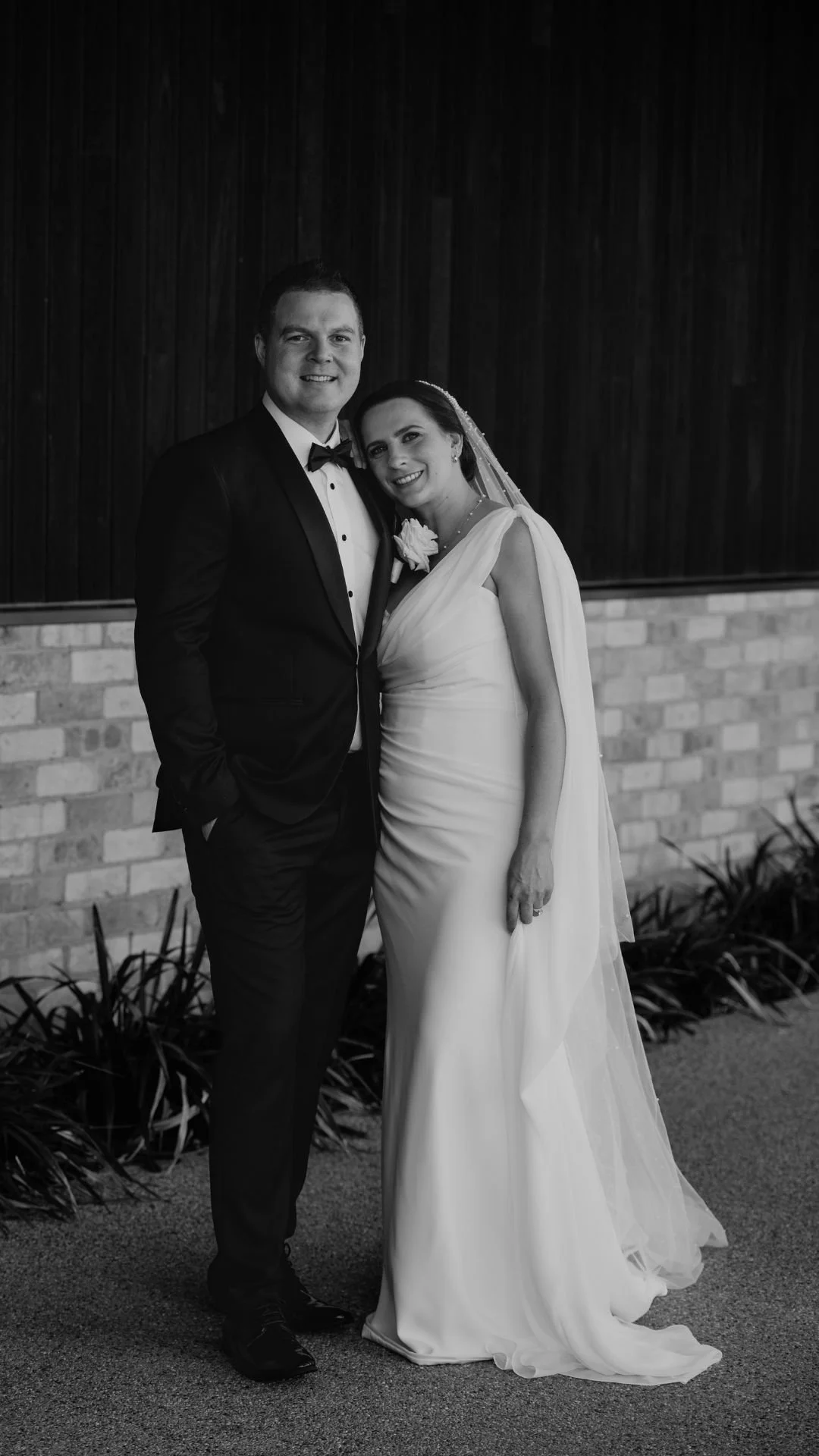 A black and white photo of a bride and groom smiling, standing together outdoors in front of a brick wall with wooden paneling, dressed in wedding attire.