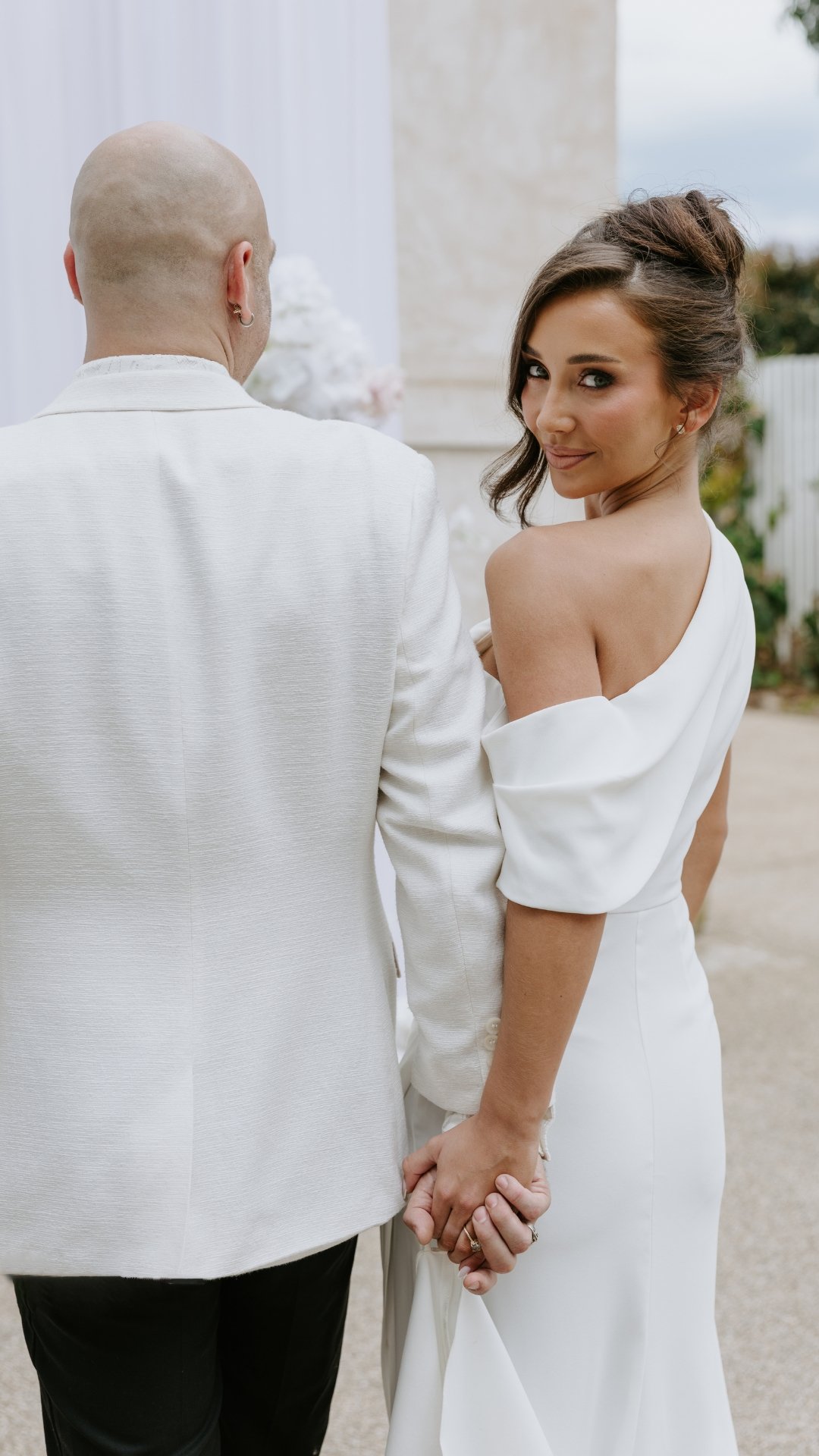 A bride and groom holding hands outdoors, with the bride smiling at the camera.