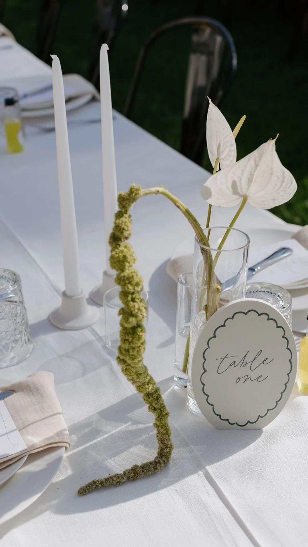 A white table setting with a clear vase holding white flowers and a green, curved decorative branch, tall white candles in candlesticks, glassware, a card labeled 'table one', and napkins.