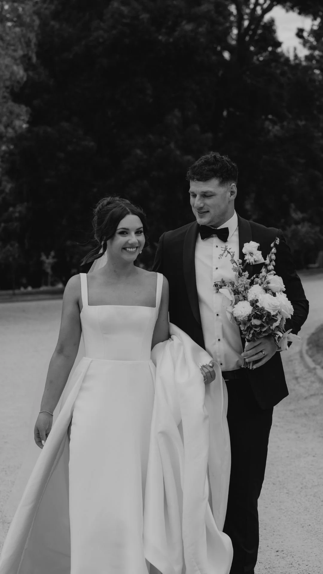 Black and white photo of a smiling bride and groom walking outdoors. The bride is wearing a sleeveless white wedding dress, and the groom is wearing a tuxedo with a bow tie, holding a bouquet of flowers.