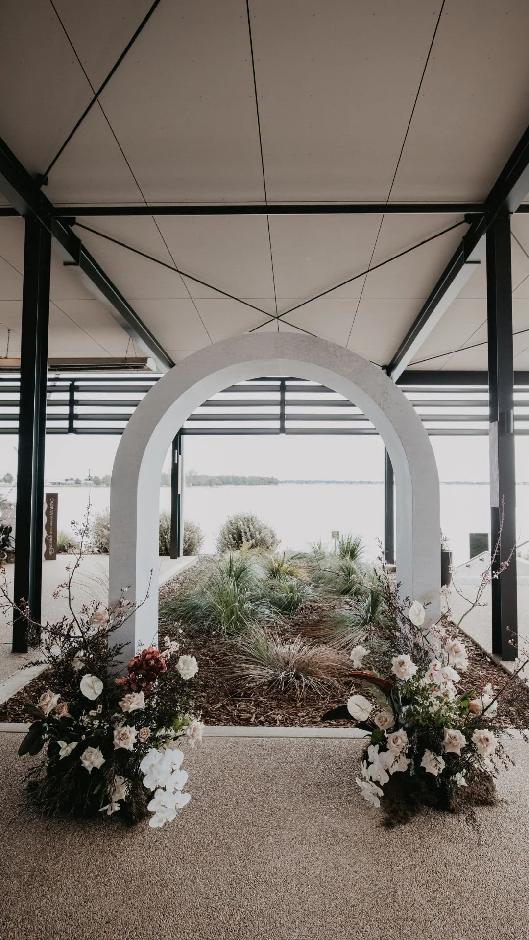 Indoor space with a garden bed filled with grasses and plants, framed by a white archway, with large windows overlooking a body of water and landscape, and decorated with floral arrangements in the foreground.