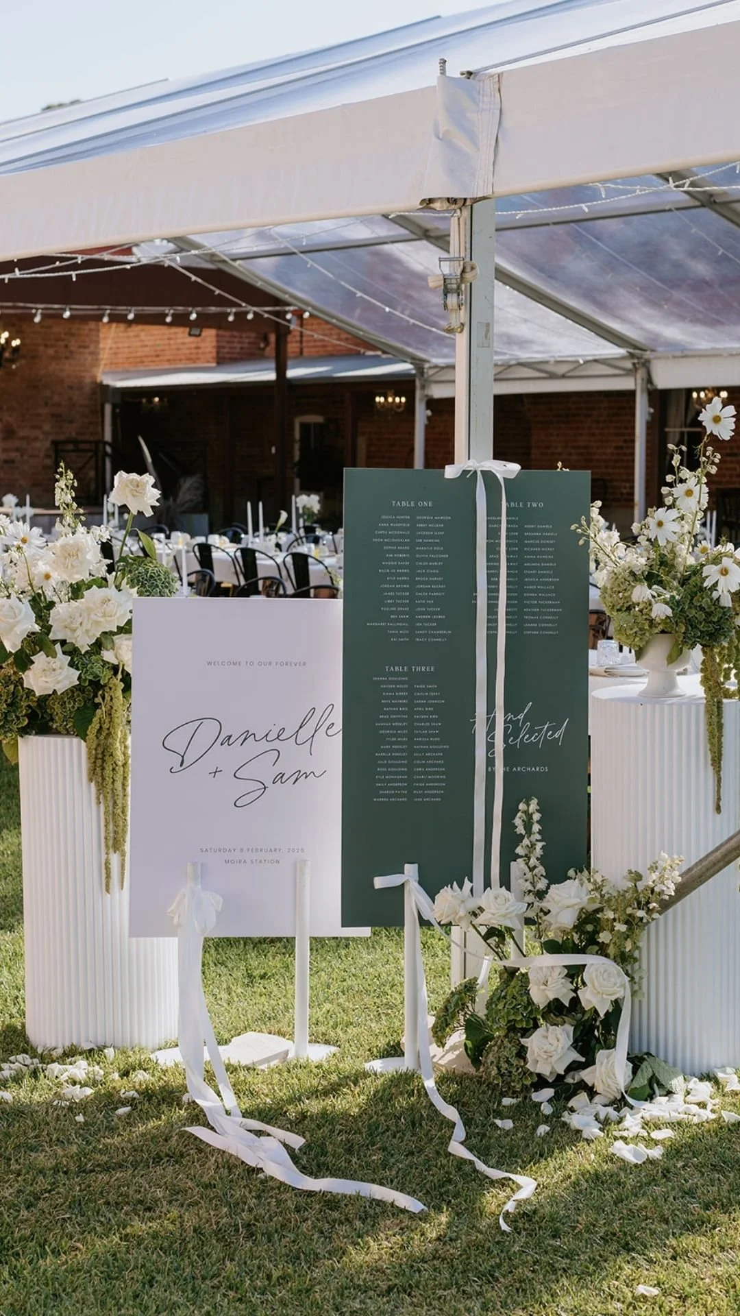 Wedding seating chart display on a white board with floral decorations at an outdoor reception. The board features the names 'Danielle + Sam' with wedding details, surrounded by white flowers and ribbon accents.