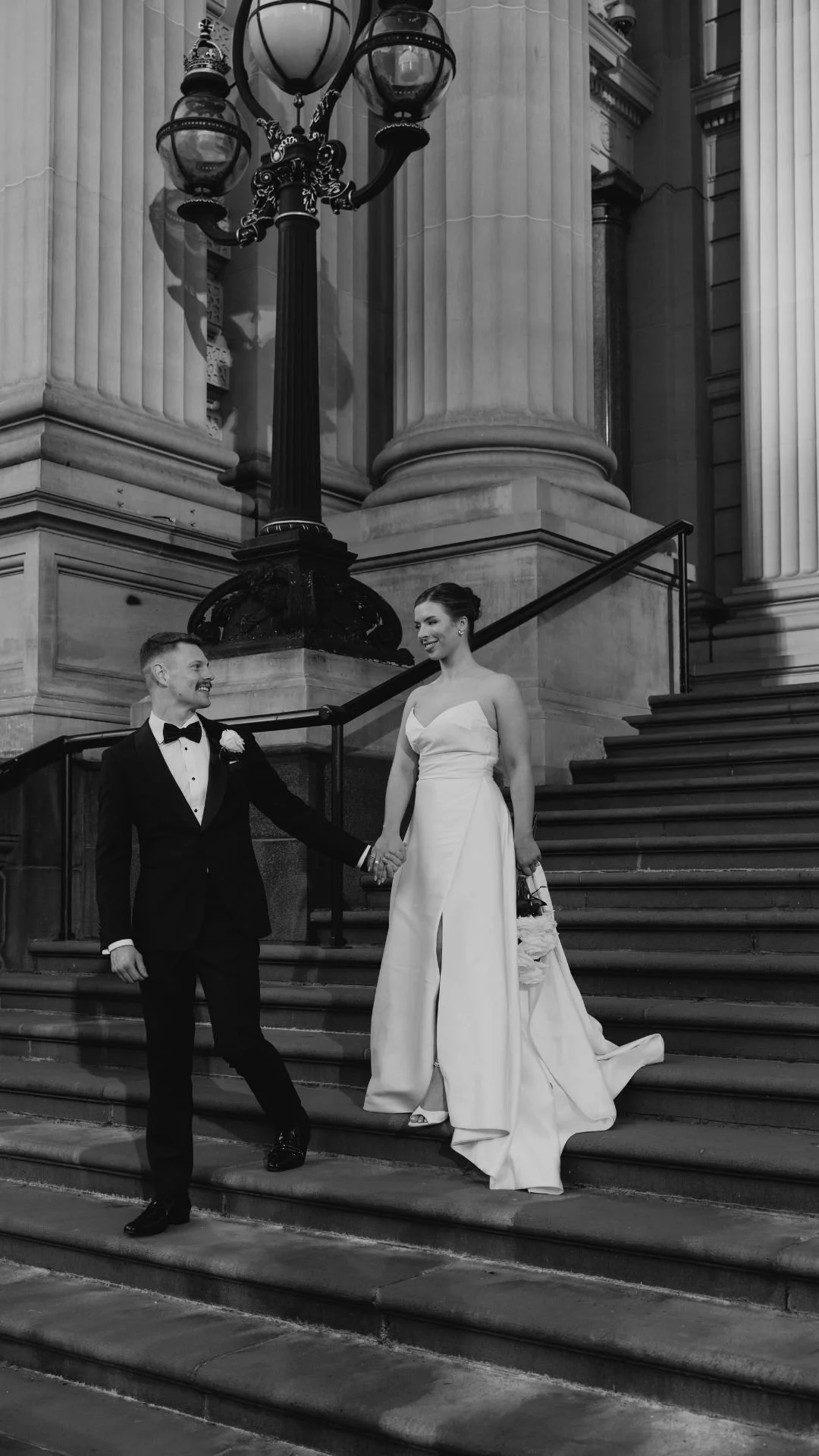 A black and white photo of a bride and groom on a staircase outside a building with large columns. The groom is in a tuxedo with a bow tie, and the bride is in a strapless wedding dress holding a bouquet, smiling at each other.