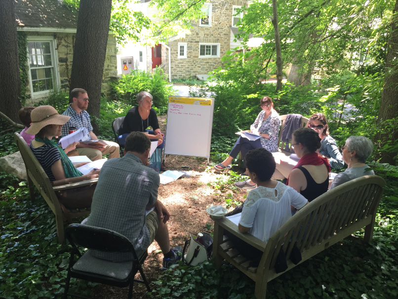 A group of people sitting in a circle outside, participating in a meeting or workshop. They are in a wooded garden area with greenery and trees. There is a flip chart with notes in the center.