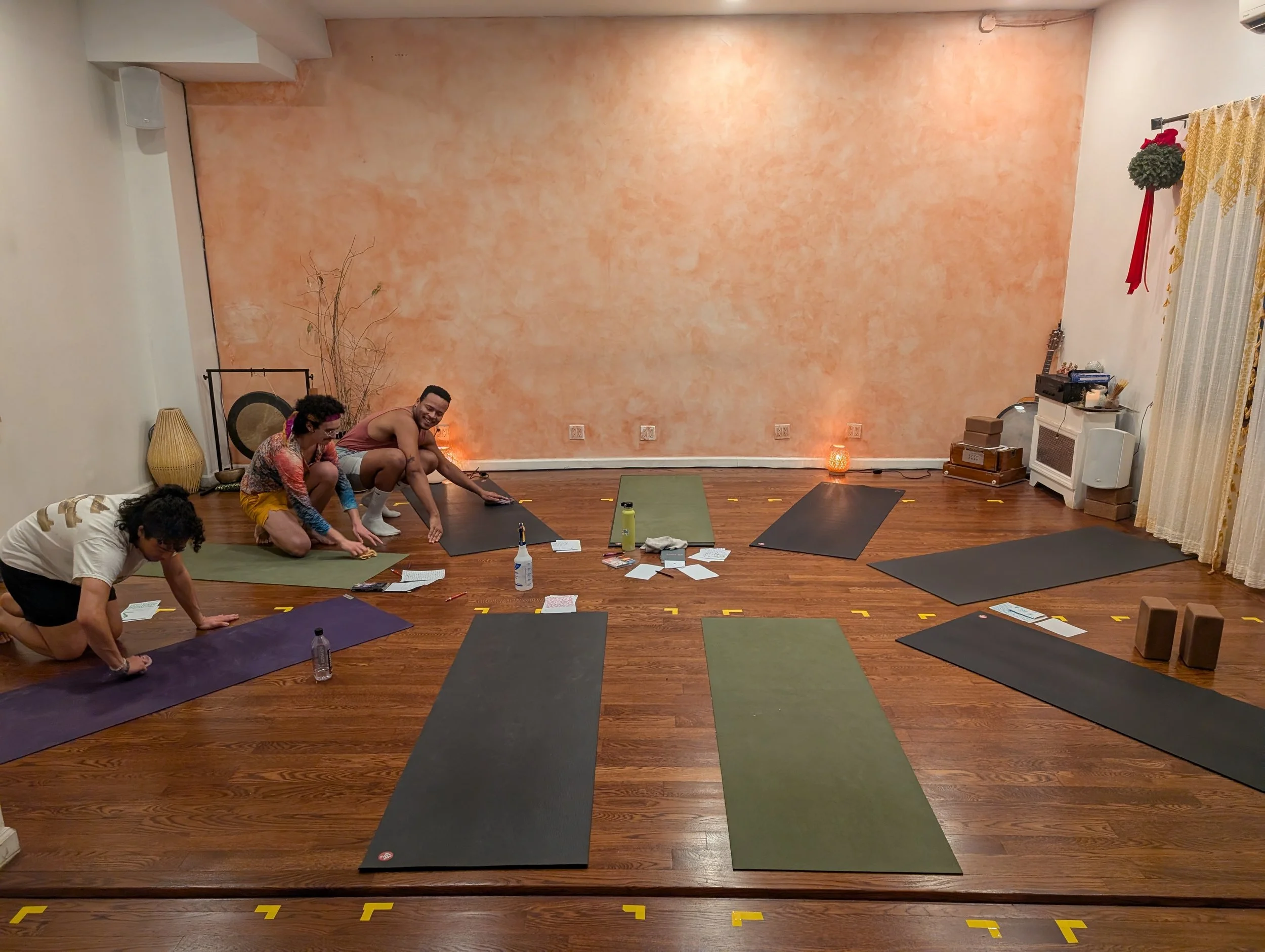 A yoga studio with six mats arranged on a wooden floor, three people preparing for a session, and decorative items including a floor lamp, a gong, and a curtain with a hanger on the wall.