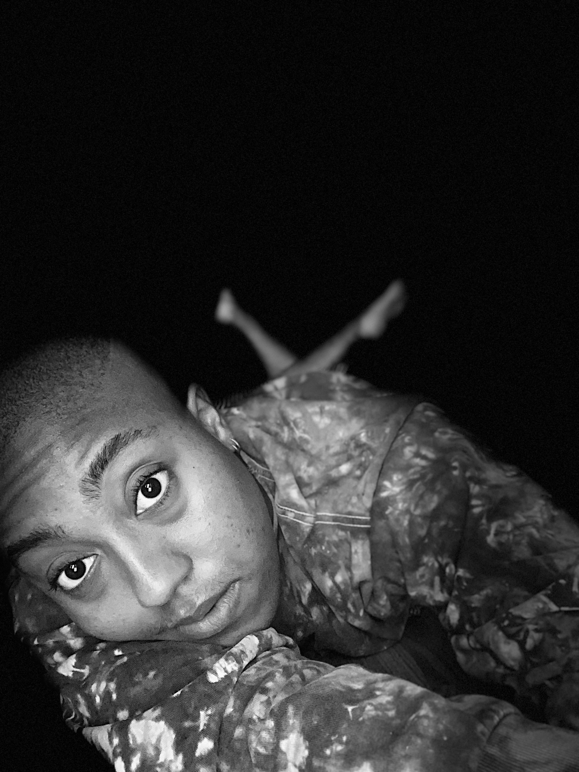 A black and white photo of a young woman with short hair, lying on her side and looking into the camera, with a dark background