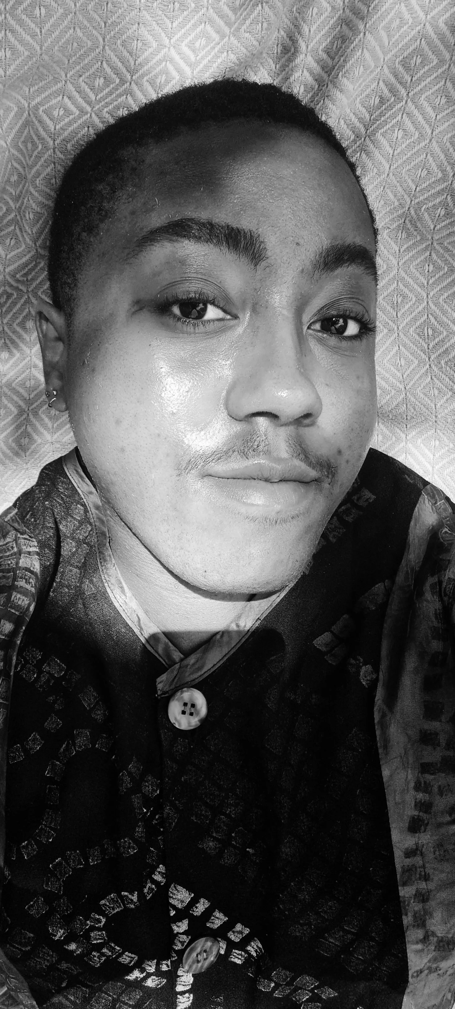 A black and white close-up photo of a young man lying on a patterned surface, with short hair, well-groomed eyebrows, and a slight smile. He's wearing earrings and a shirt with large buttons and a textured pattern.