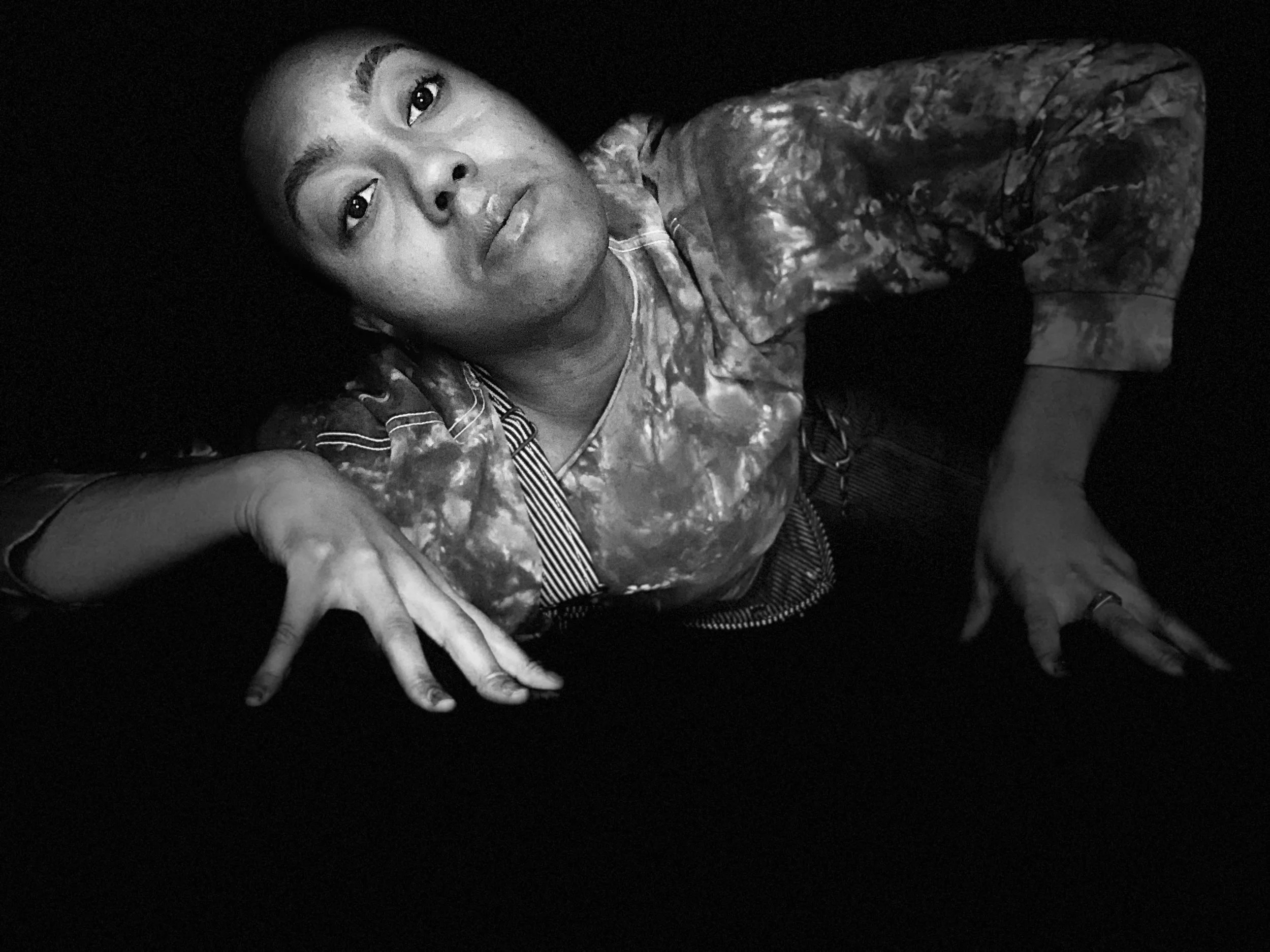 A nonbinary person with a tie-dye long-sleeve shirt lying on their side with his head propped up, looking directly at the camera. They have short hair, and the photo is in black and white.