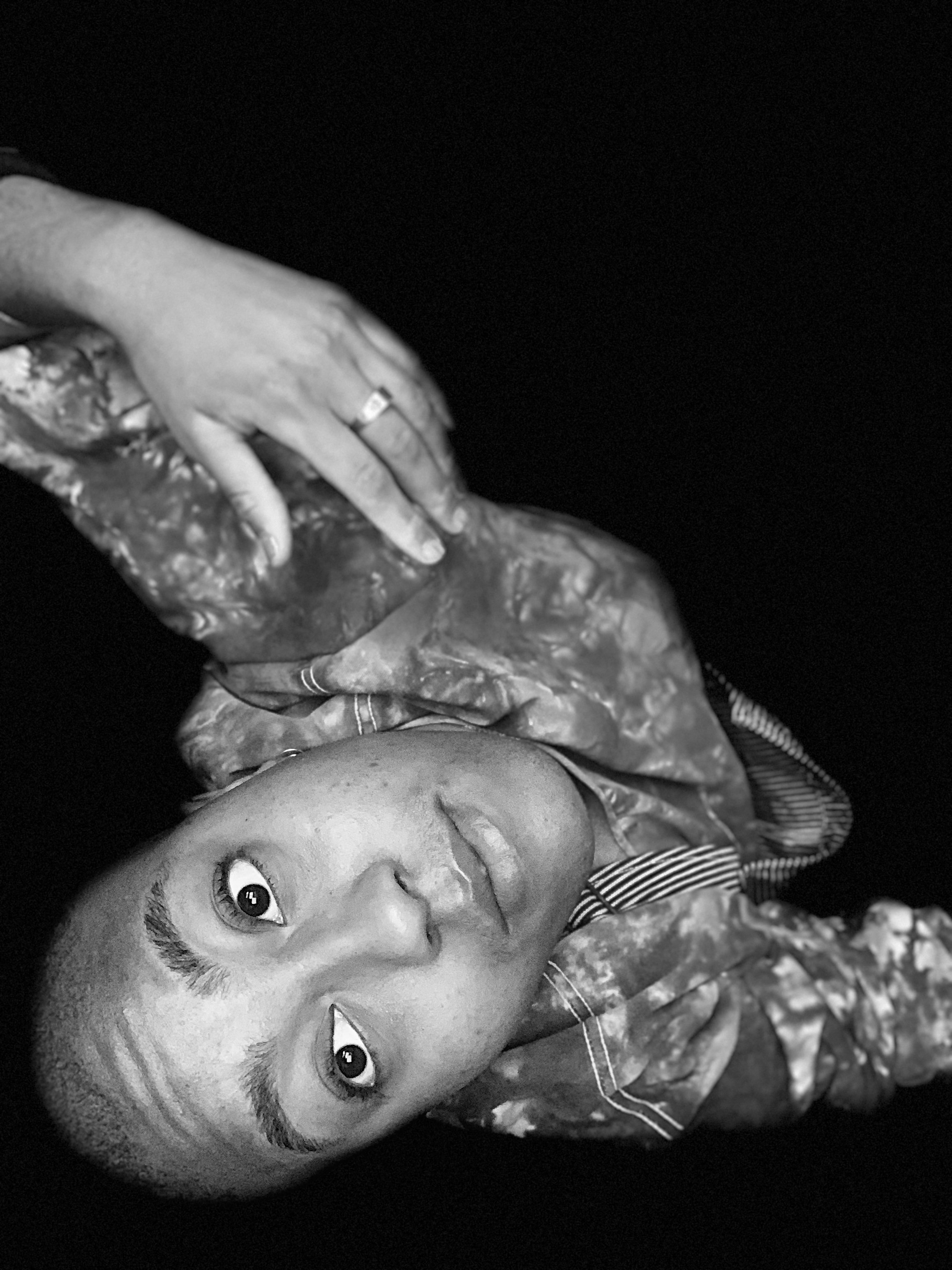 A person poses with their head resting on their hand, wearing a tie-dye shirt, looking directly at the camera with an inquisitive expression, in black and white.