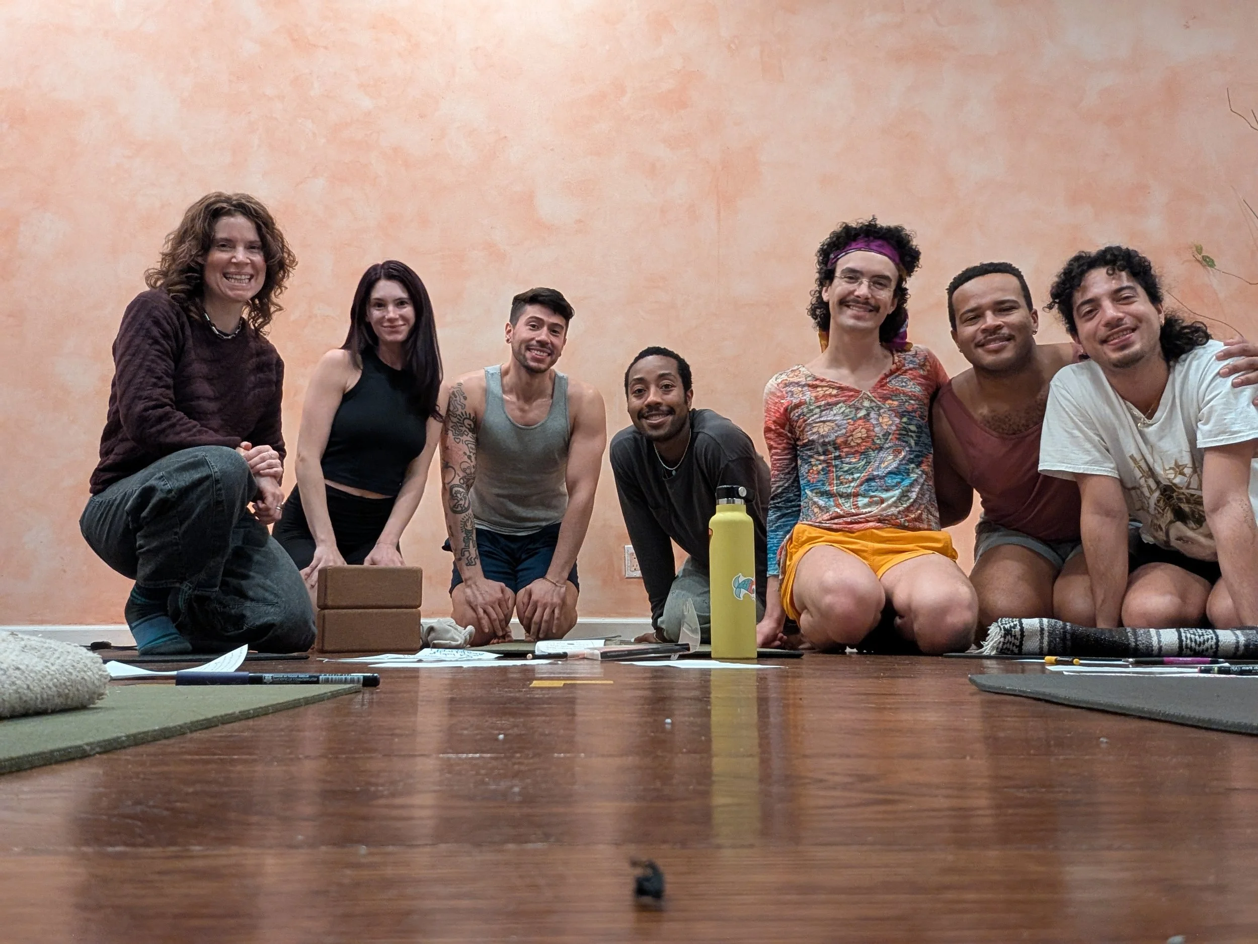 A group of eight people sitting on their knees on a wooden floor, smiling at the camera, in front of a plain peach-colored wall, with yoga mats and water bottles nearby.