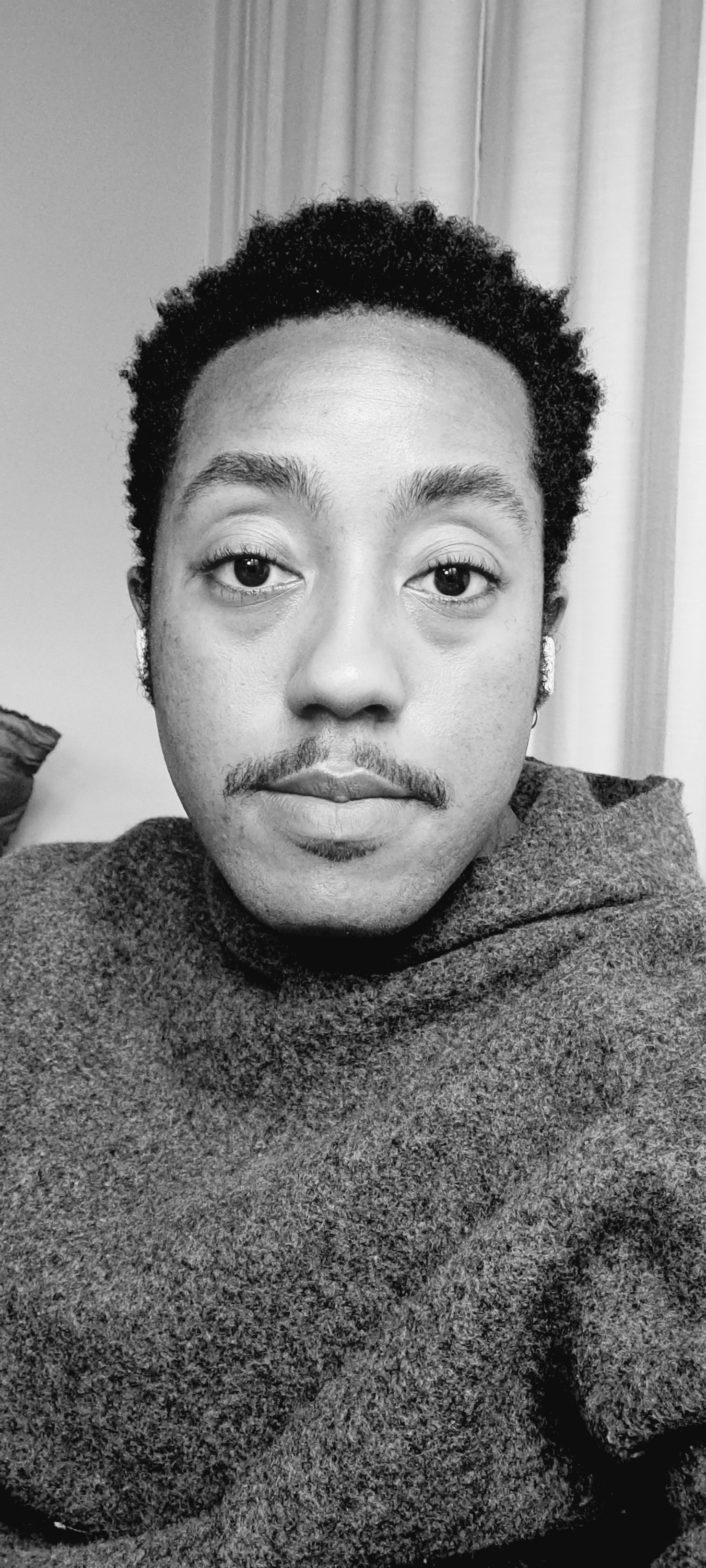 Black and white photo of a person with short curly hair, wearing earrings and a textured sweater, sitting indoors near a curtain.