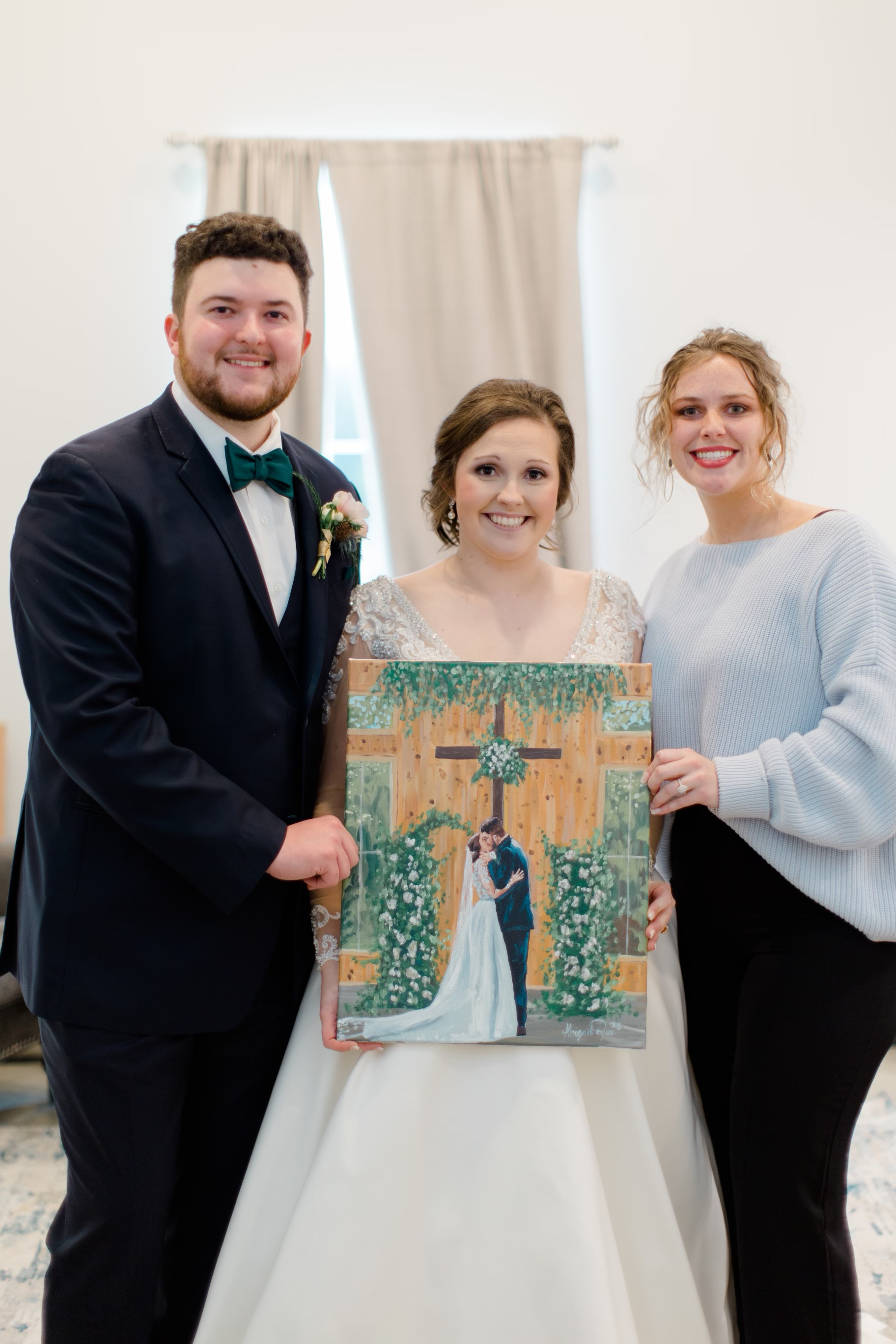 Three people, including a bride in a white dress, groom in a dark suit, and woman in a light sweater, holding a wedding painting. The painting depicts a couple at a wedding ceremony with floral decorations.