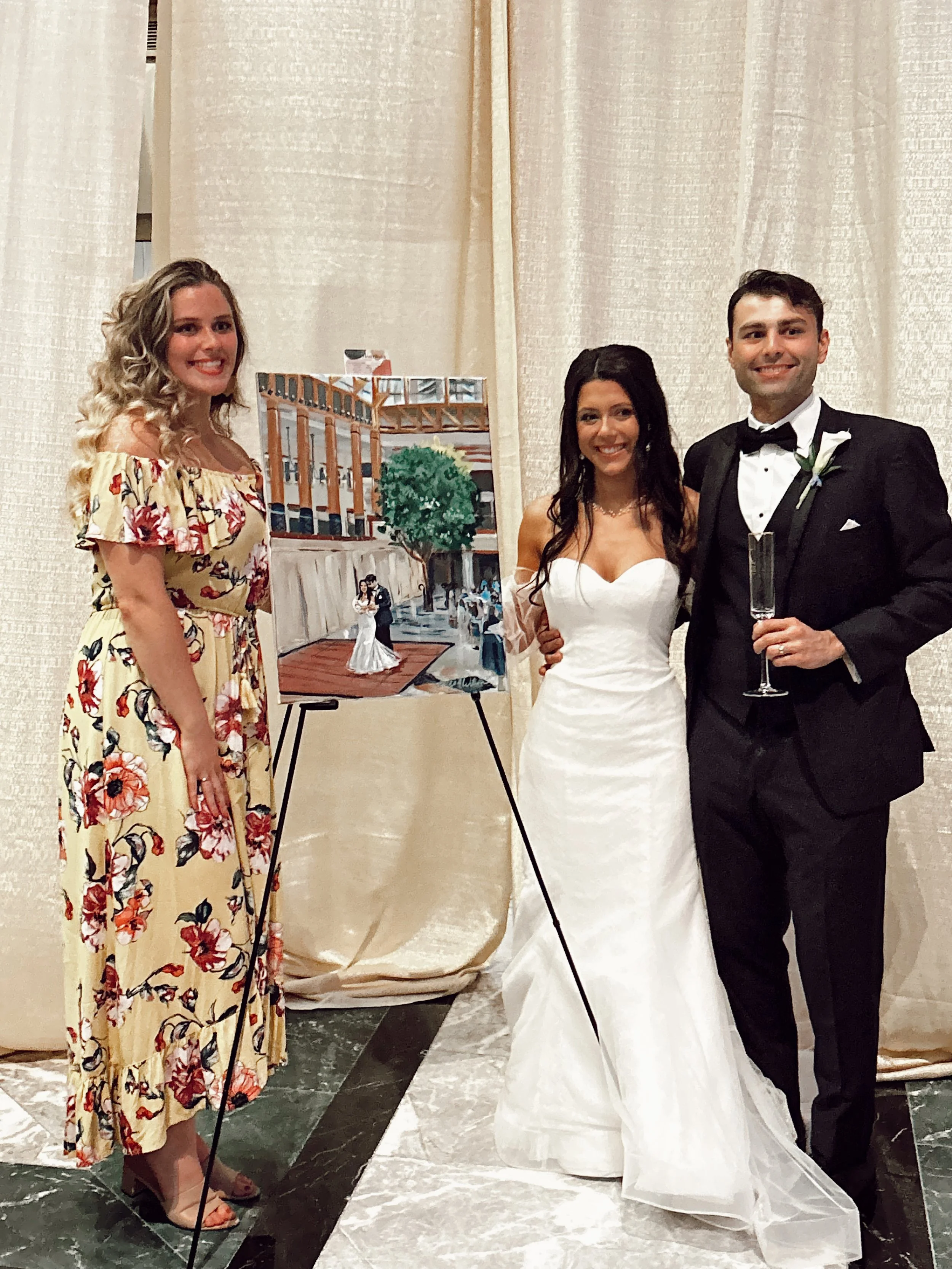 Wedding couple and a guest posing with a painted portrait on an easel. The bride is in a strapless white gown, the groom in a tuxedo, and the guest in a floral off-the-shoulder dress.