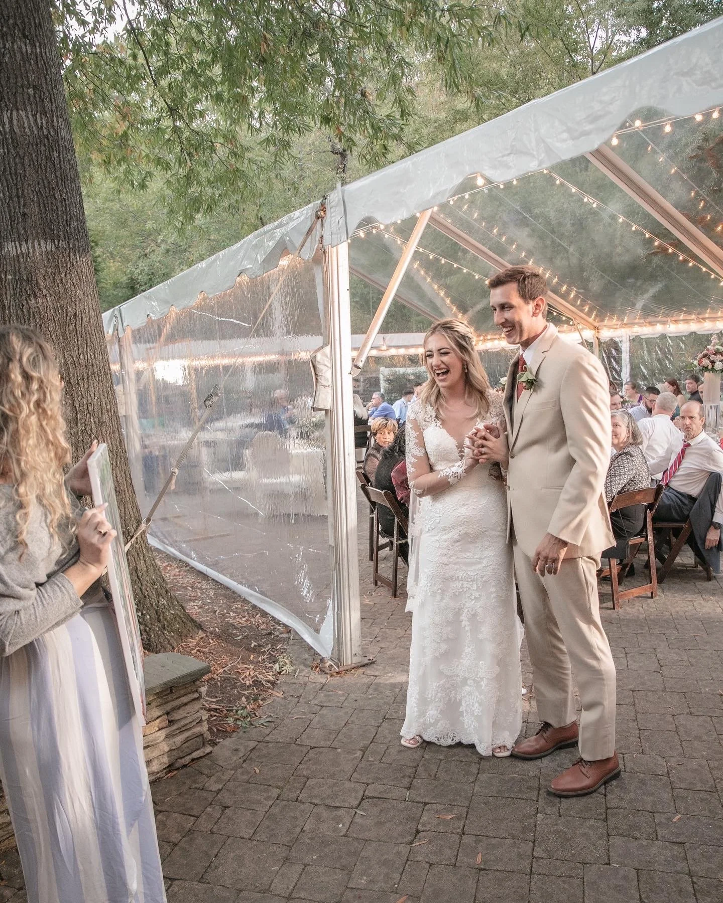 A bride and groom smiling at their wedding reception under a clear tent with lights. A woman with curly hair stands to the side holding a board, while guests are seated in the background.