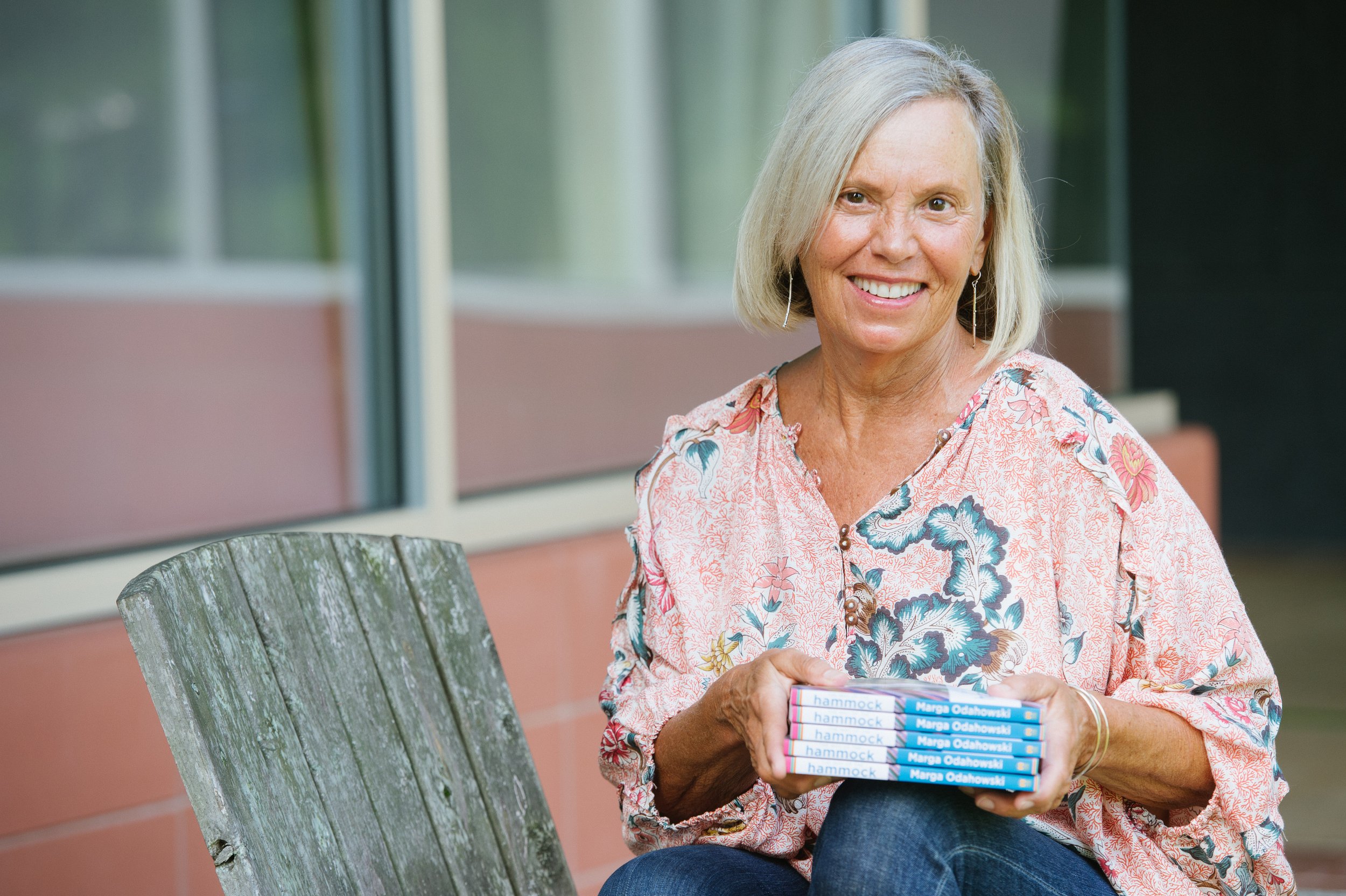 An elderly woman with blonde hair smiling while holding a stack of books titled "Hammock" by Marge Odahowski, sitting outdoors in front of a pink building with large windows.